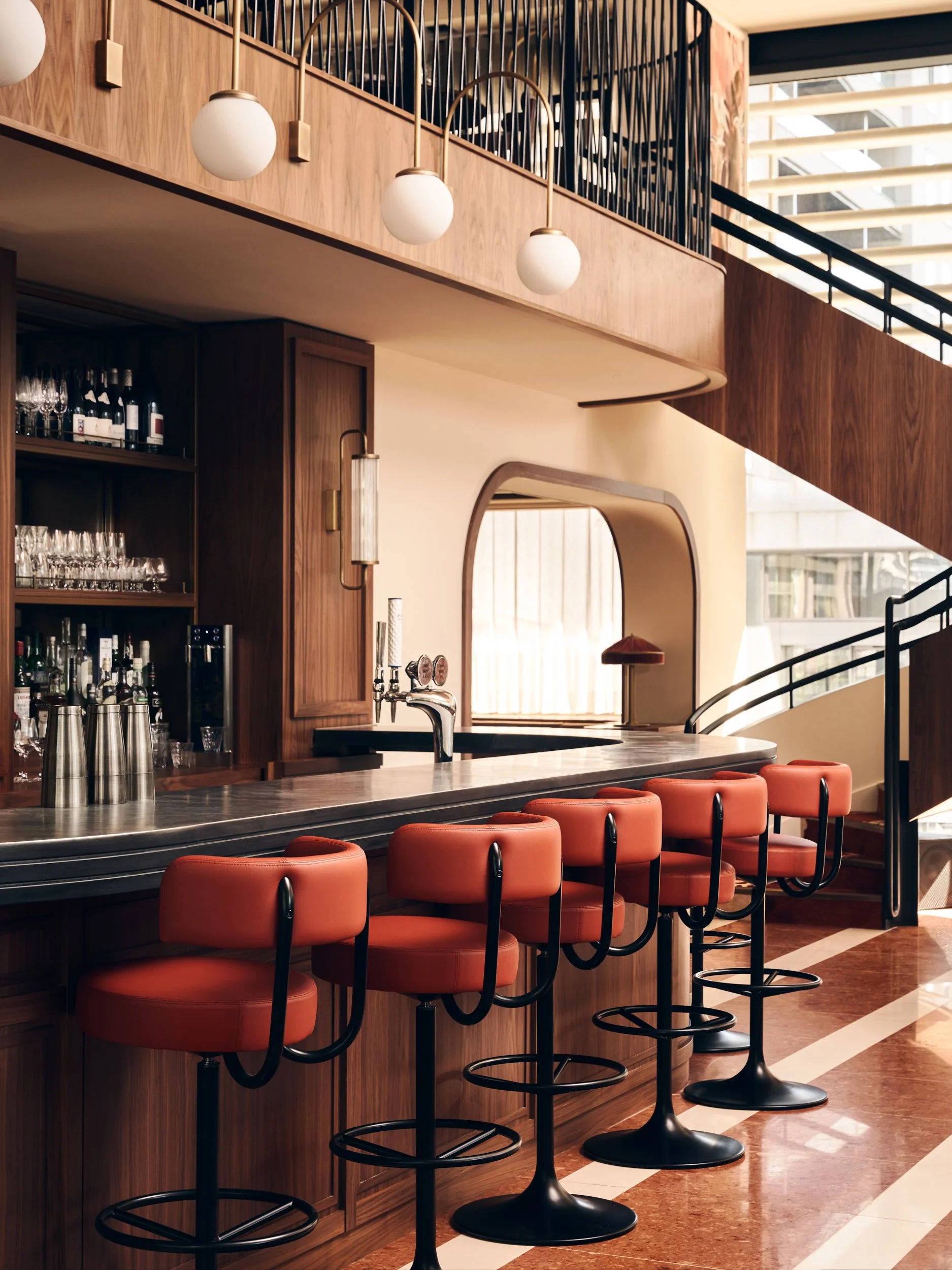 Interior of a modern bar with a wooden counter, orange bar stools, and a shelving area with glasses and bottles, featuring mid-century lighting and design elements.