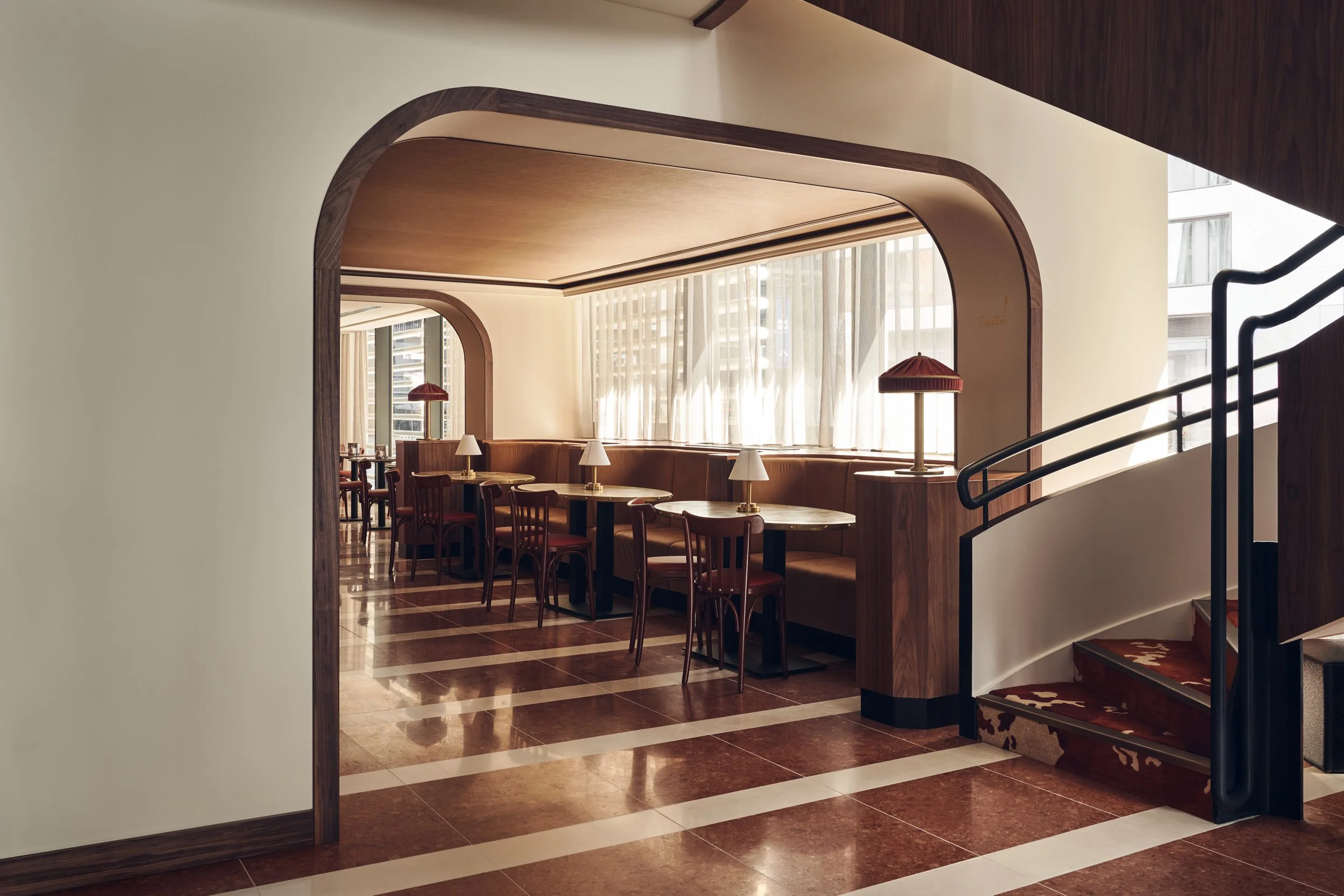 Interior view of a modern restaurant with arched wooden accents, tables with chairs and lamps, large windows with curtains, and a staircase with black handrails.
