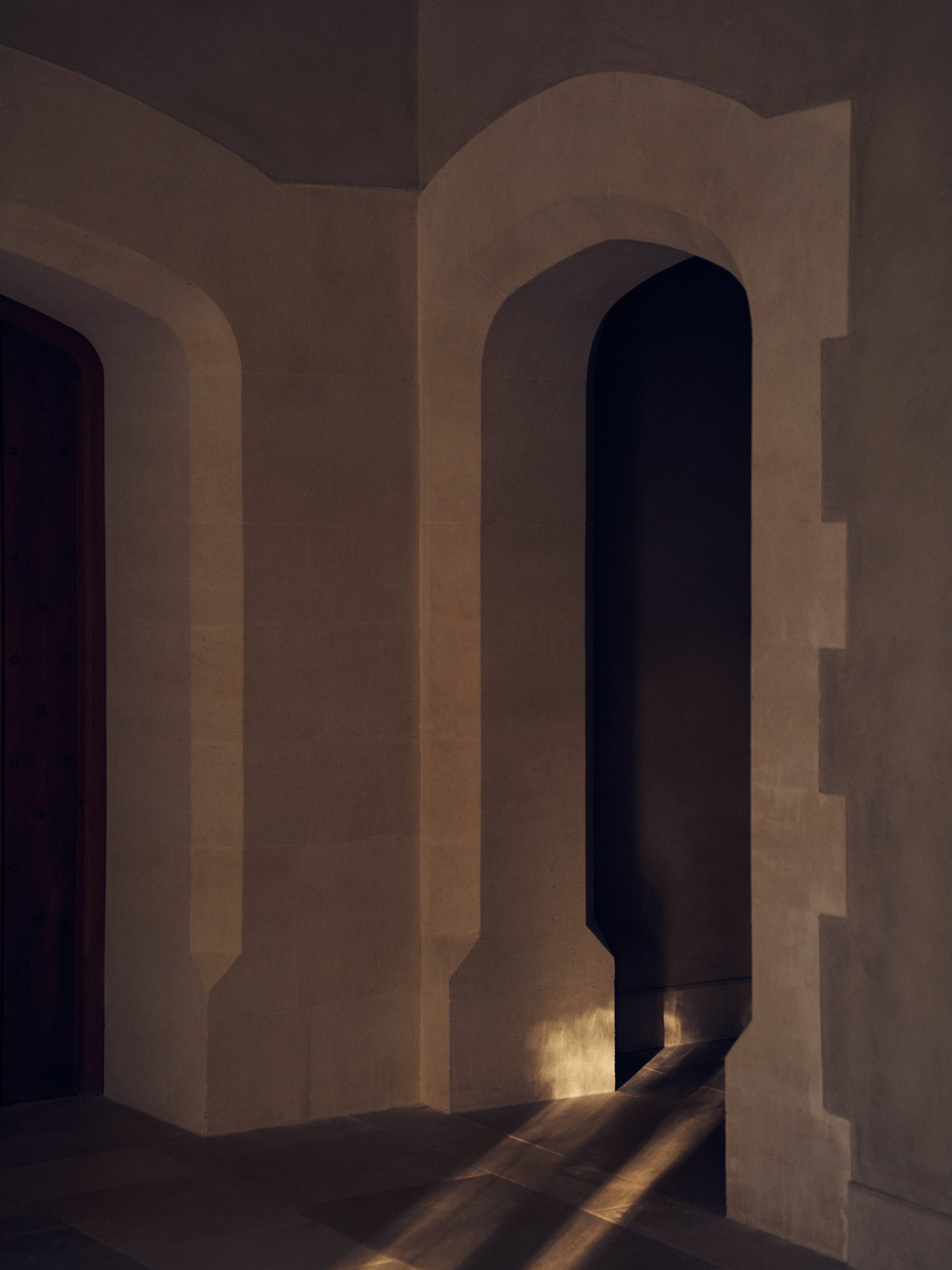 A dimly lit corridor with beige stone walls featuring arches and staircases