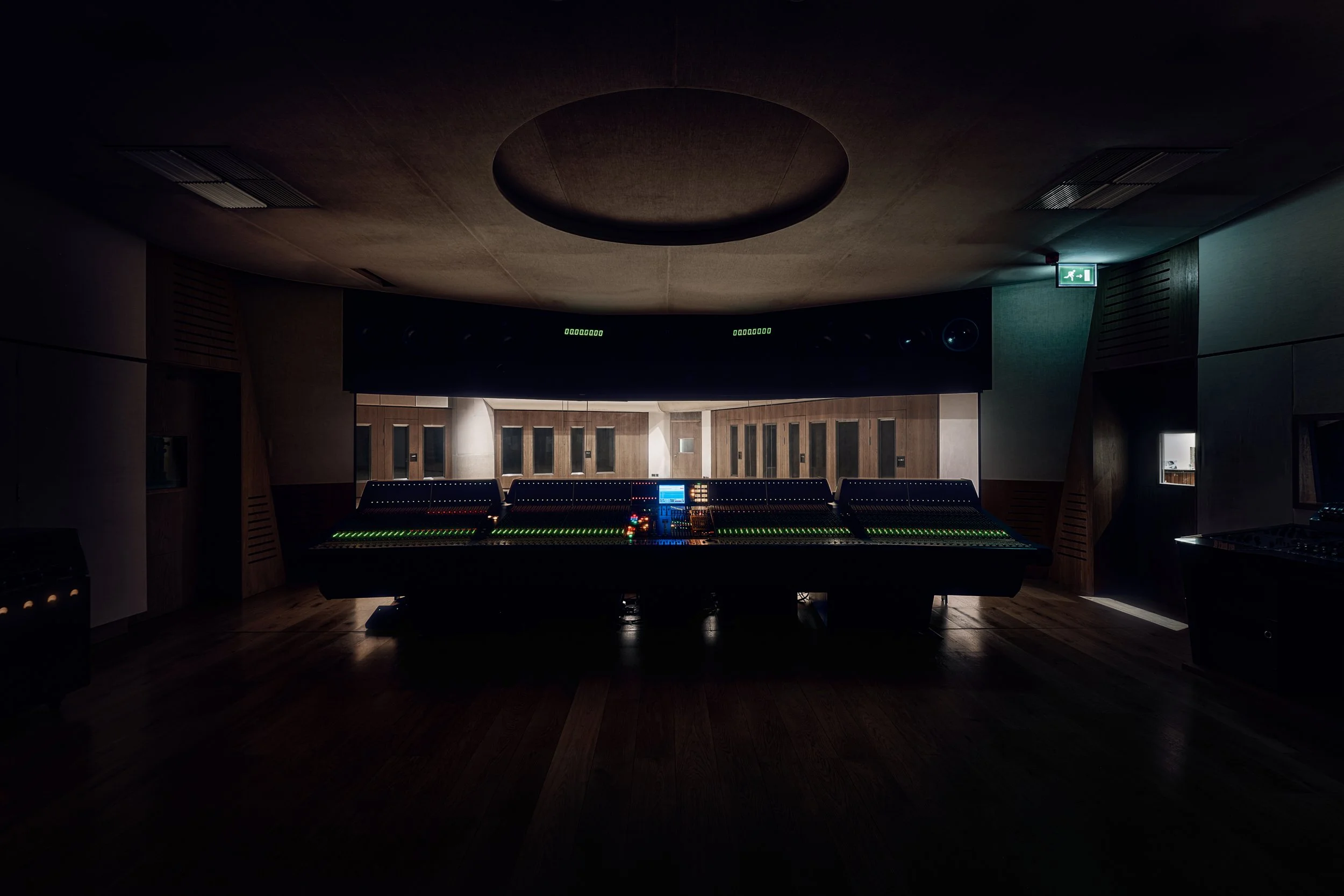 A dimly lit recording studio control room with a large mixing console featuring numerous faders and colorful lights, and a dark ceiling with a circular indentation.