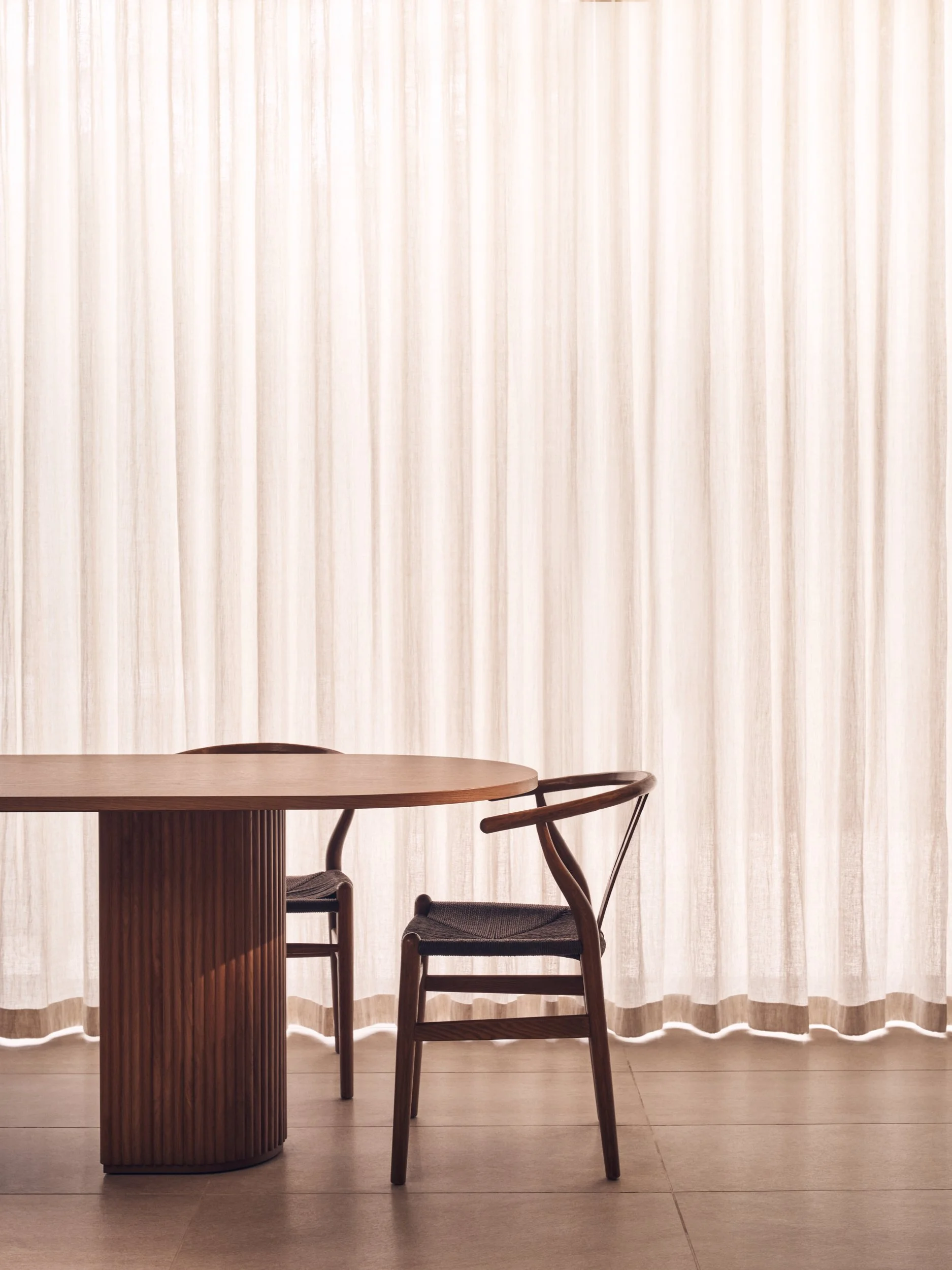A wooden table with a rounded edge, accompanied by two chairs with woven seats, positioned in front of a beige, semi-transparent curtain with vertical stripes.