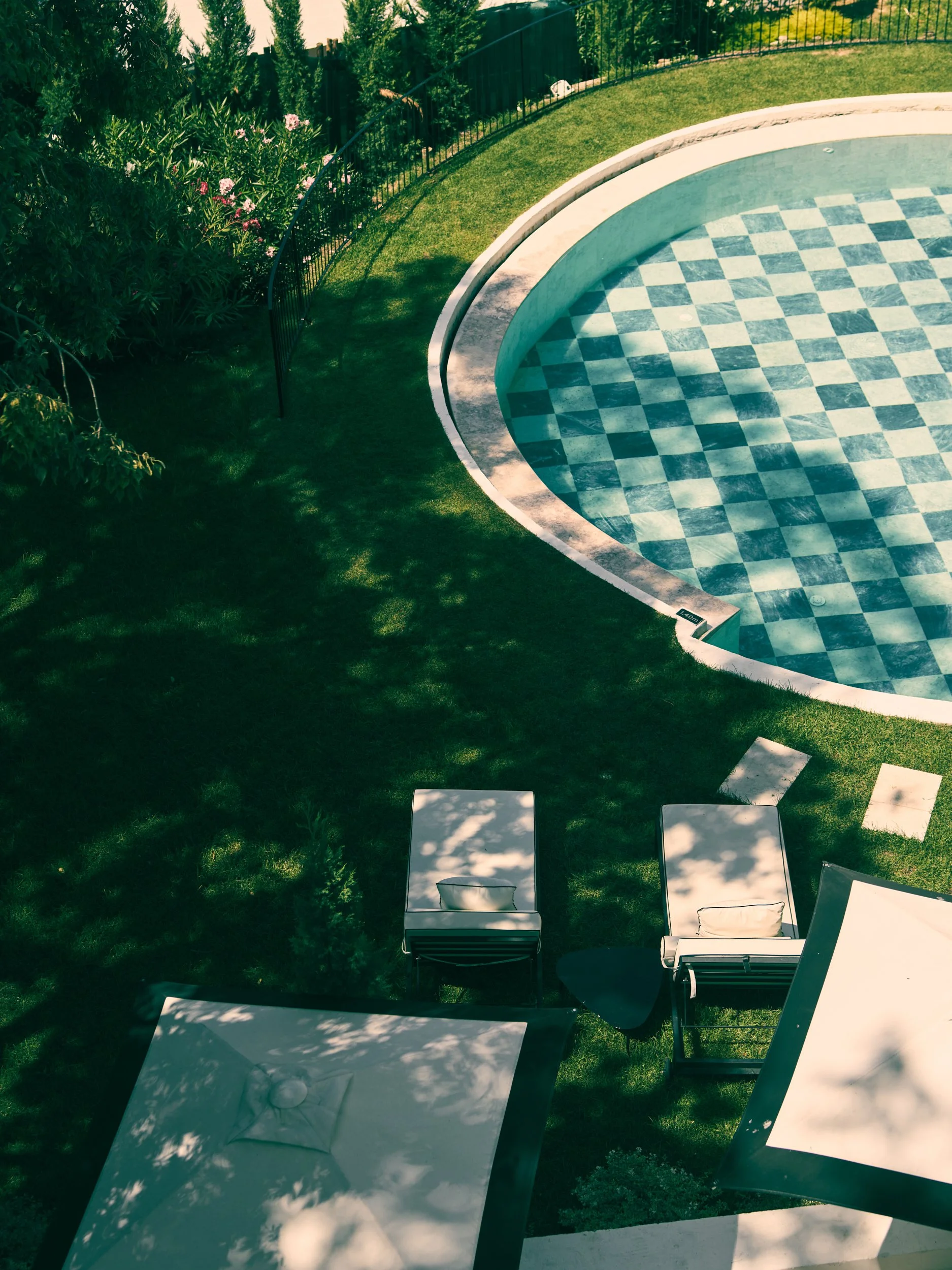 Overshot view of a backyard with a swimming pool, surrounded by green grass, trees, and a black fence.