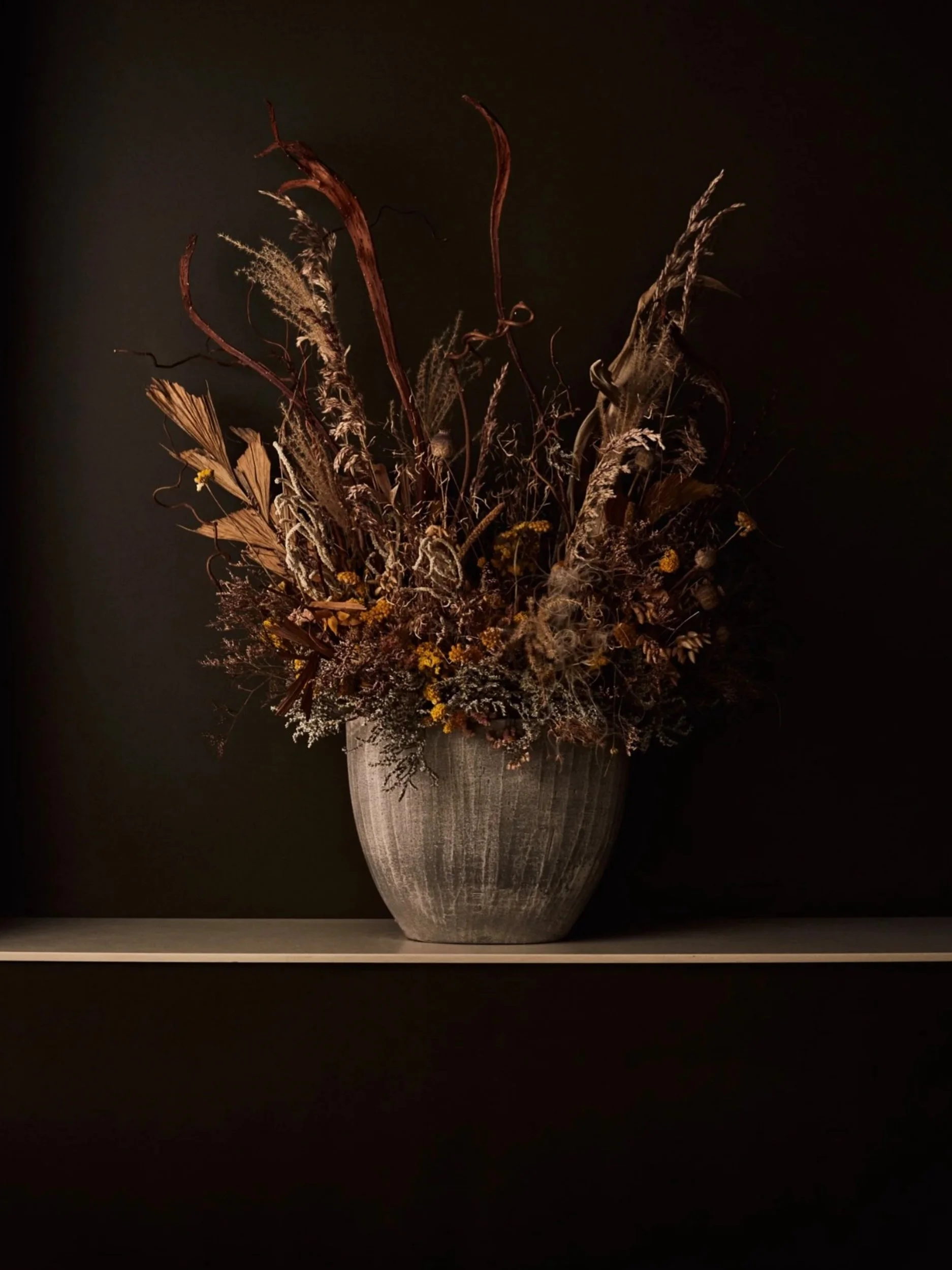 A tall, textured, gray ceramic vase filled with dried flowers and grasses, set against a black background.