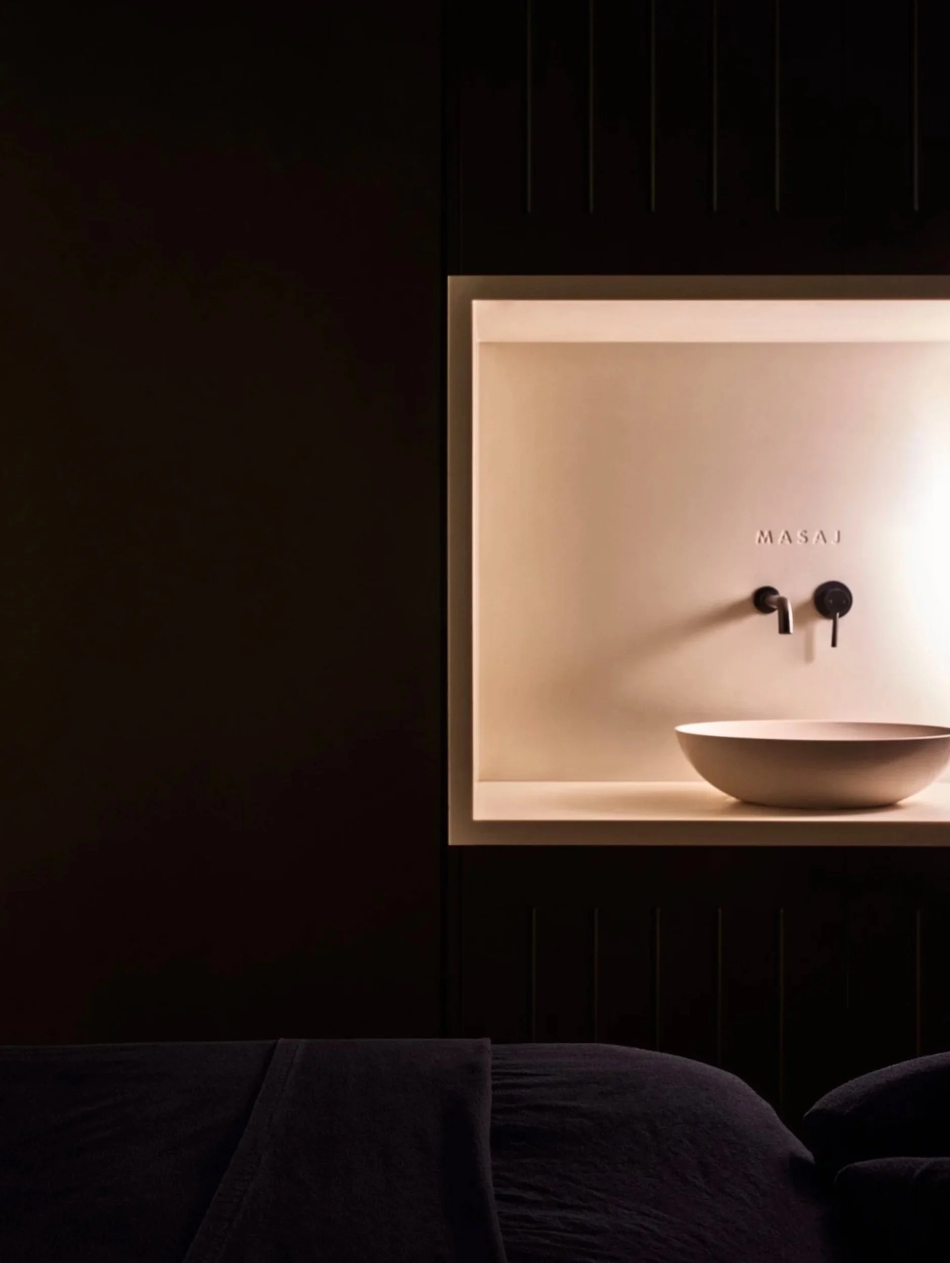 A dimly lit room with a bed in the foreground and a wall-mounted specifically designed sink with the brand name 'MASAJ' visible, along with two faucets, set against a beige wall.
