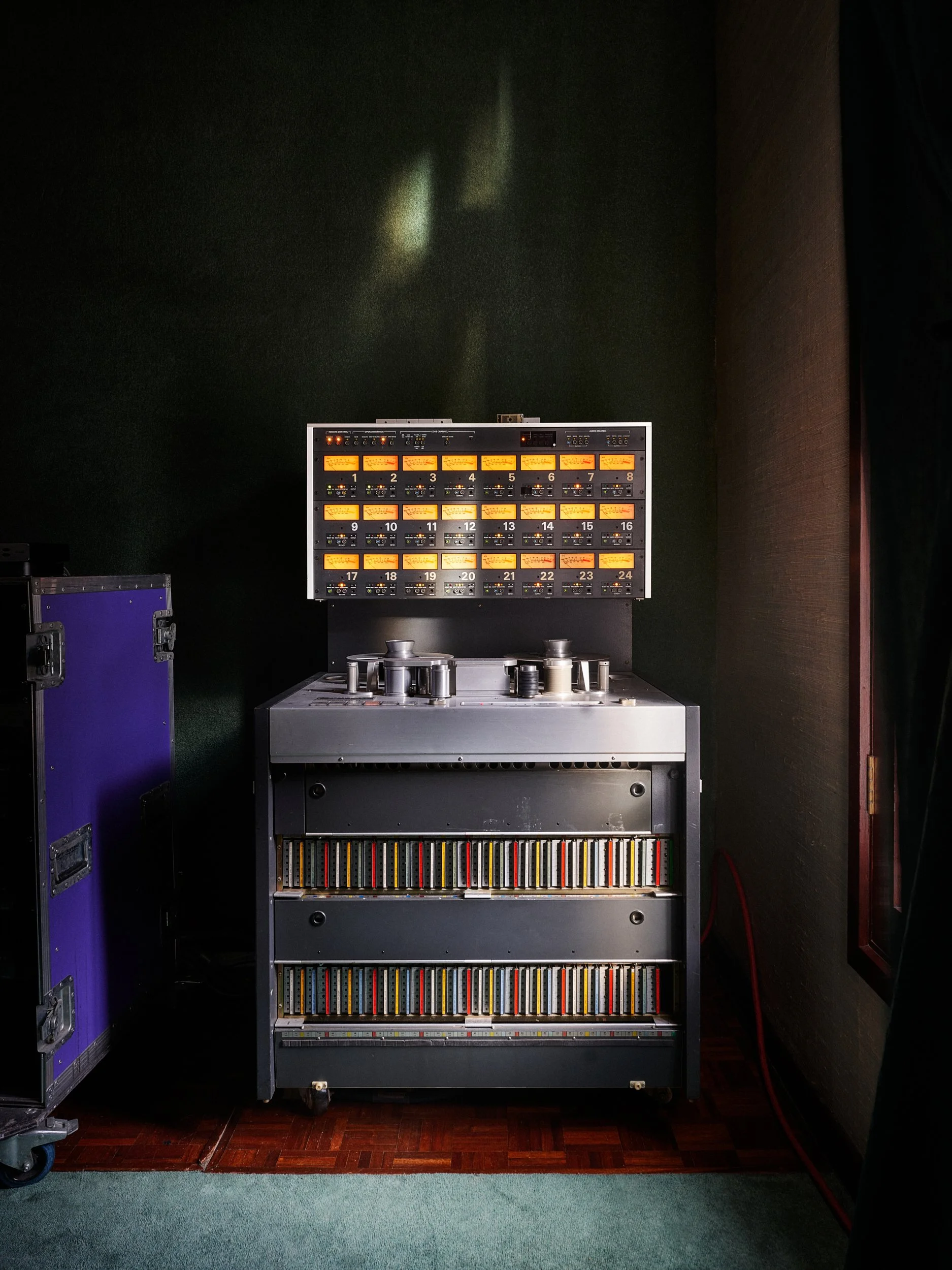 A vintage audio mixing console with numerous illuminated yellow and orange meters and control knobs, placed on a black stand against a dark green wall, with what appears to be audio equipment and cassette tapes stored below.