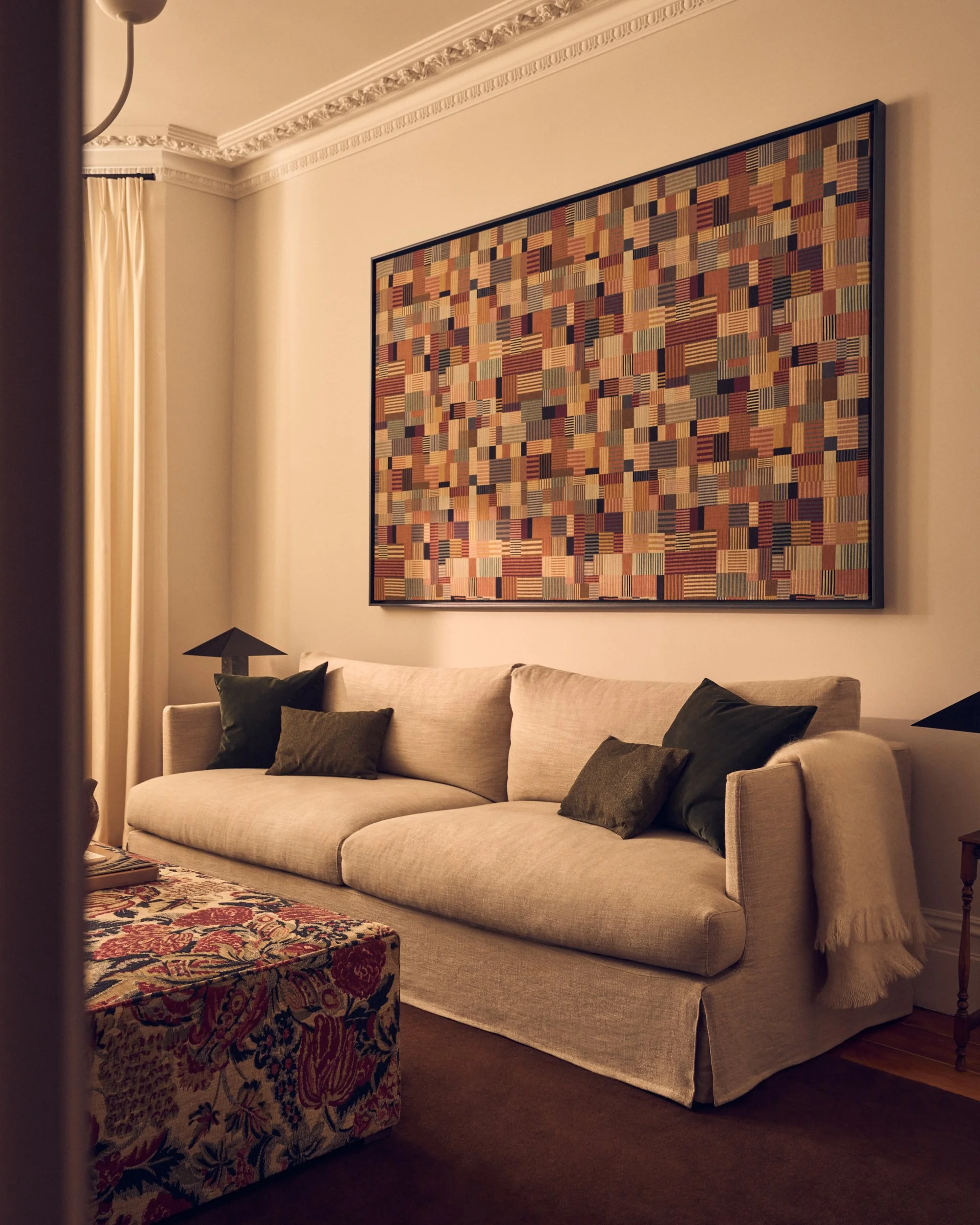 A living room corner with a beige couch adorned with dark green and beige pillows, a part of a rug with floral patterns, a side table with a dark lampshade, a large colorful abstract artwork on the wall, and cream-colored curtains by the window.