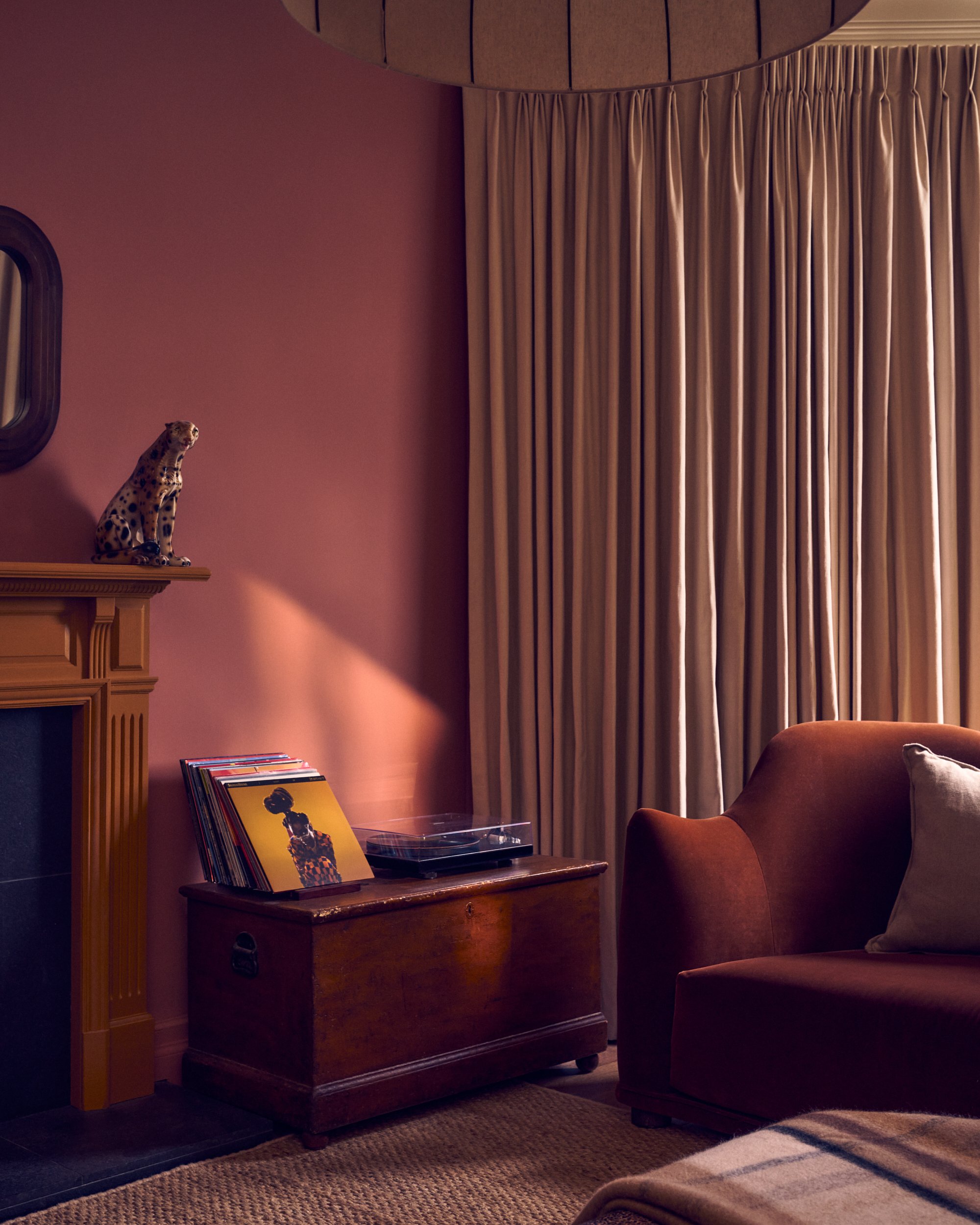 A living room corner with a wooden credenza, a stack of vinyl records, a record player, beige curtains, a brown armchair, and a small mirror on the wall.