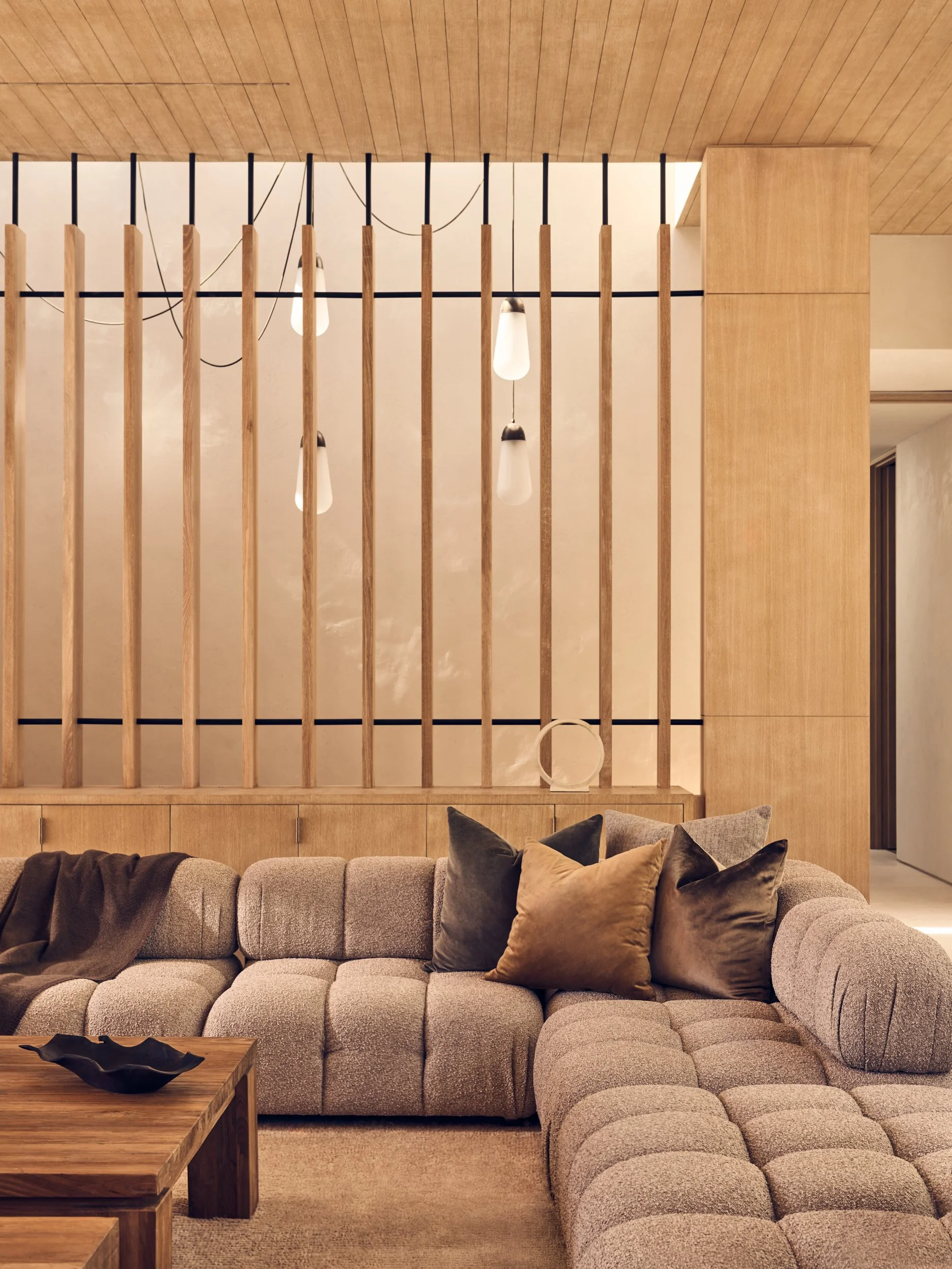A cozy living room with a beige, plush sectional sofa decorated with black, brown, and beige pillows, in front of a wooden partition with vertical slats and hanging light fixtures. A wooden coffee table with a black decorative bowl is in the foregrou