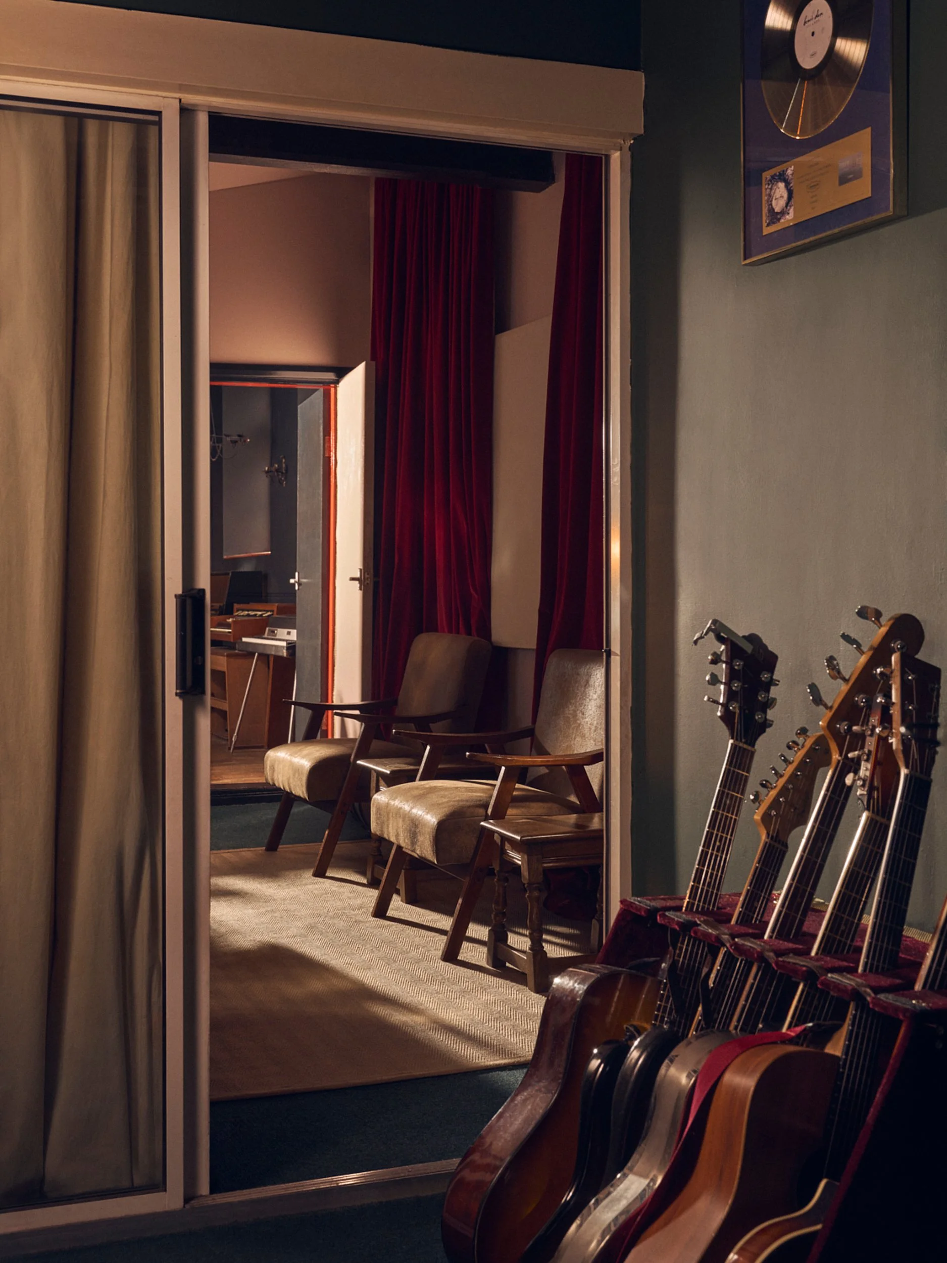 A recording studio with a wall framed gold record. Inside, there are three wooden chairs with upholstered seats, a carpeted floor, and a large red velvet curtain. Several guitars rest against the wall near the chairs.