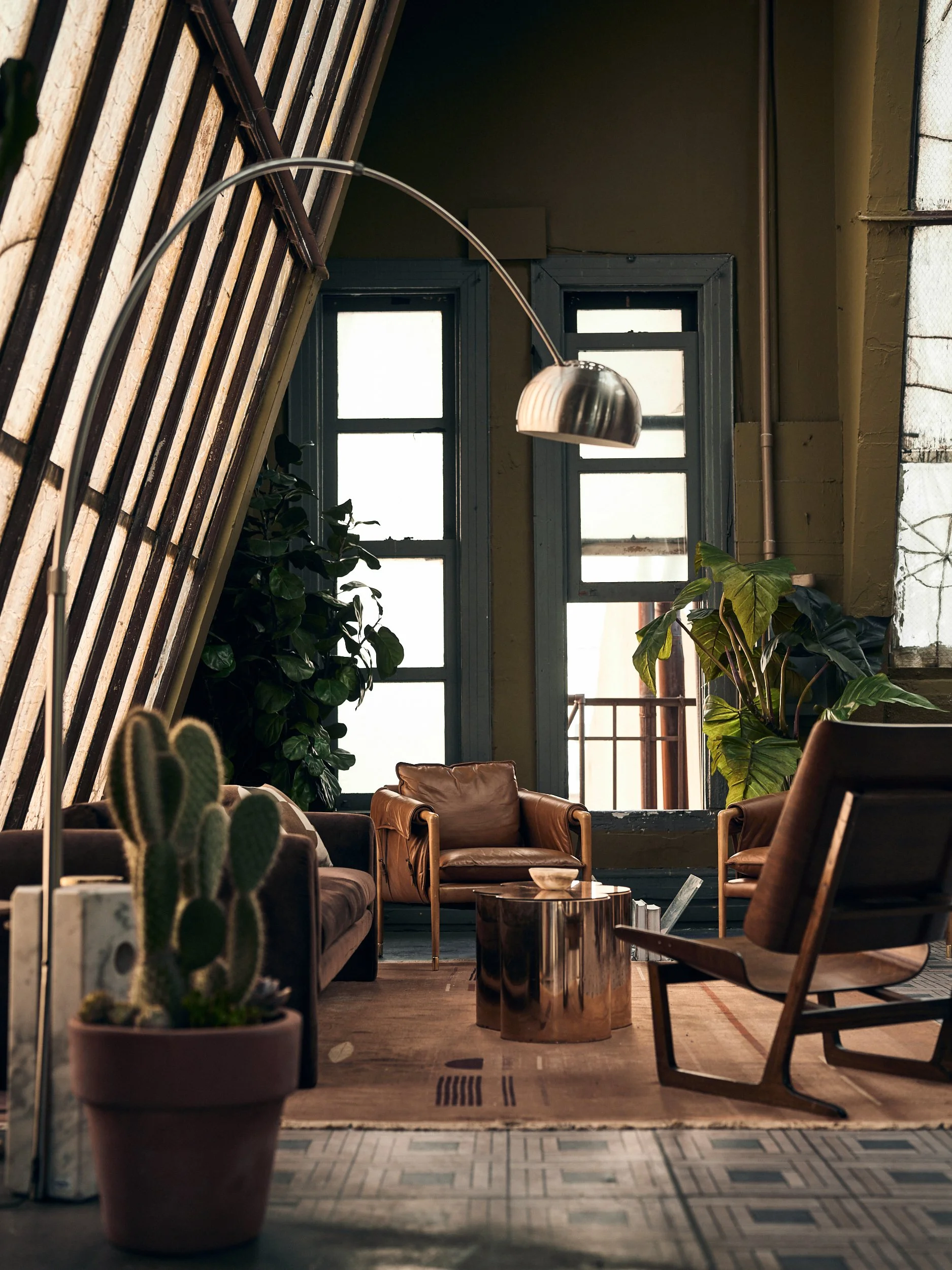 Cozy loft-style living room with large windows, indoor plants, leather armchairs, a wooden coffee table, and modern decor.
