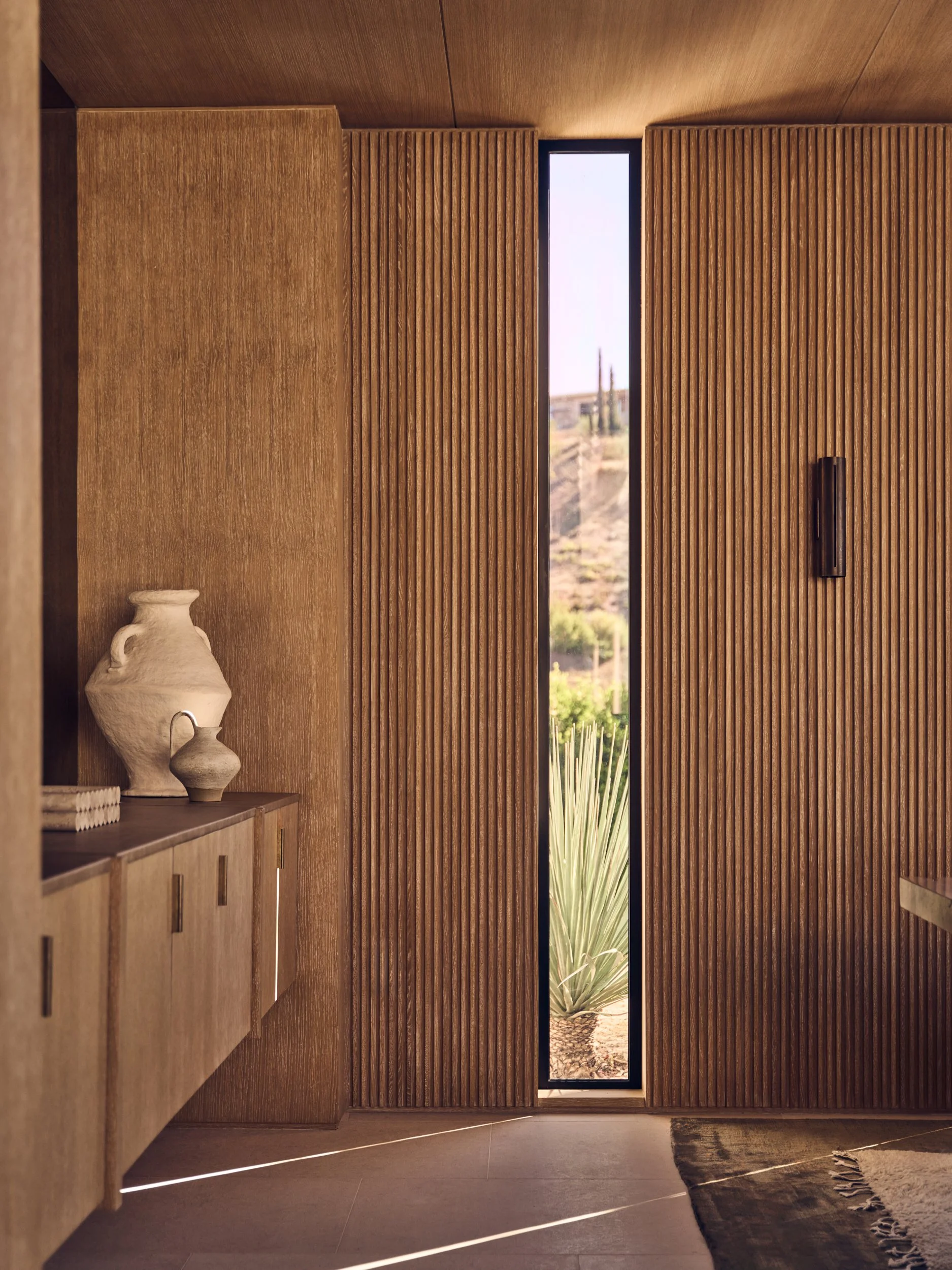 Interior view of a modern entryway with wooden sliding doors, a narrow vertical window showing an outdoor landscape, a sideboard with decorative vases, and a rug on the floor.