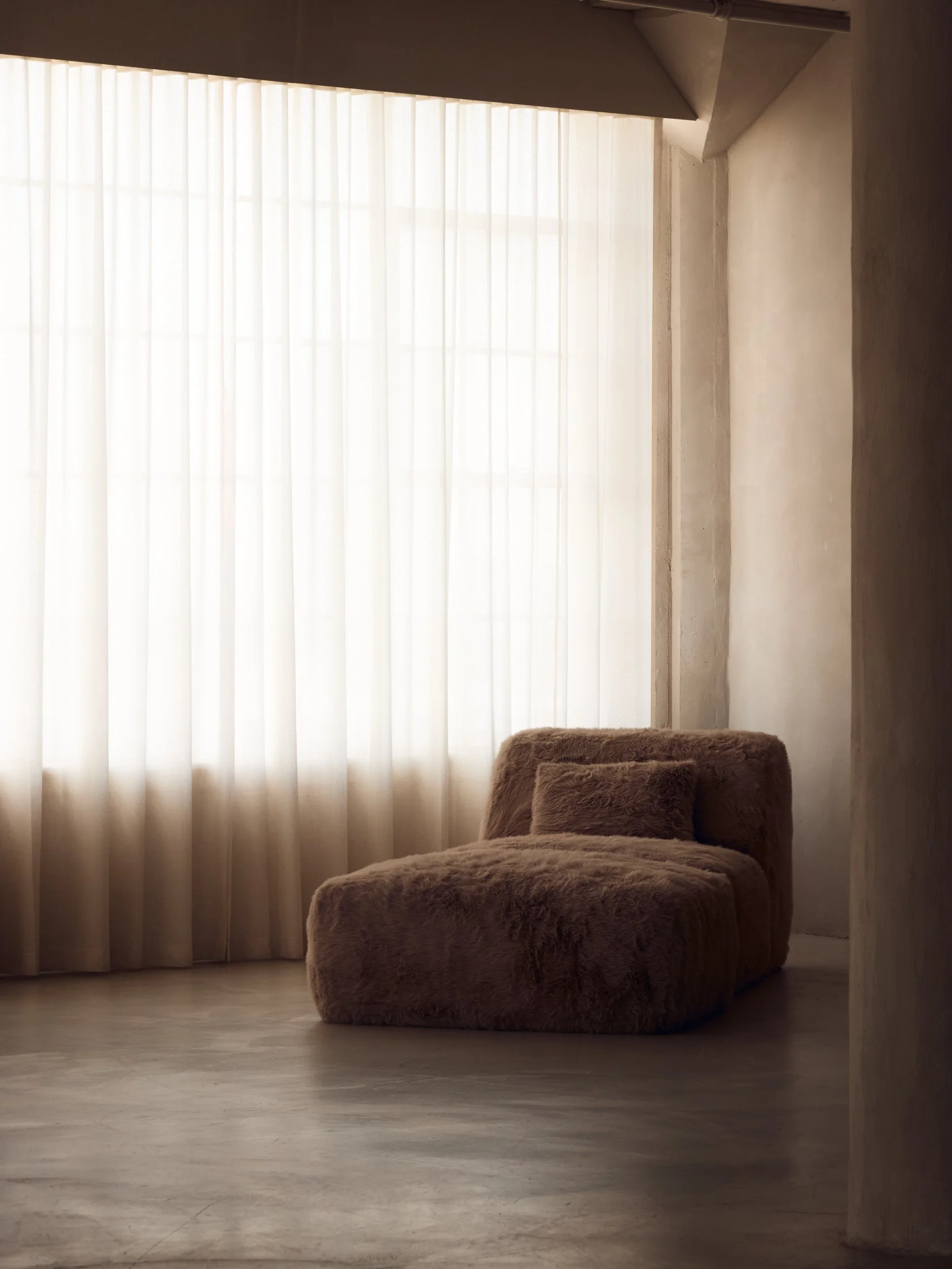 A plush beige armchair and a matching ottoman in front of a large window with sheer white curtains, in a softly lit room.