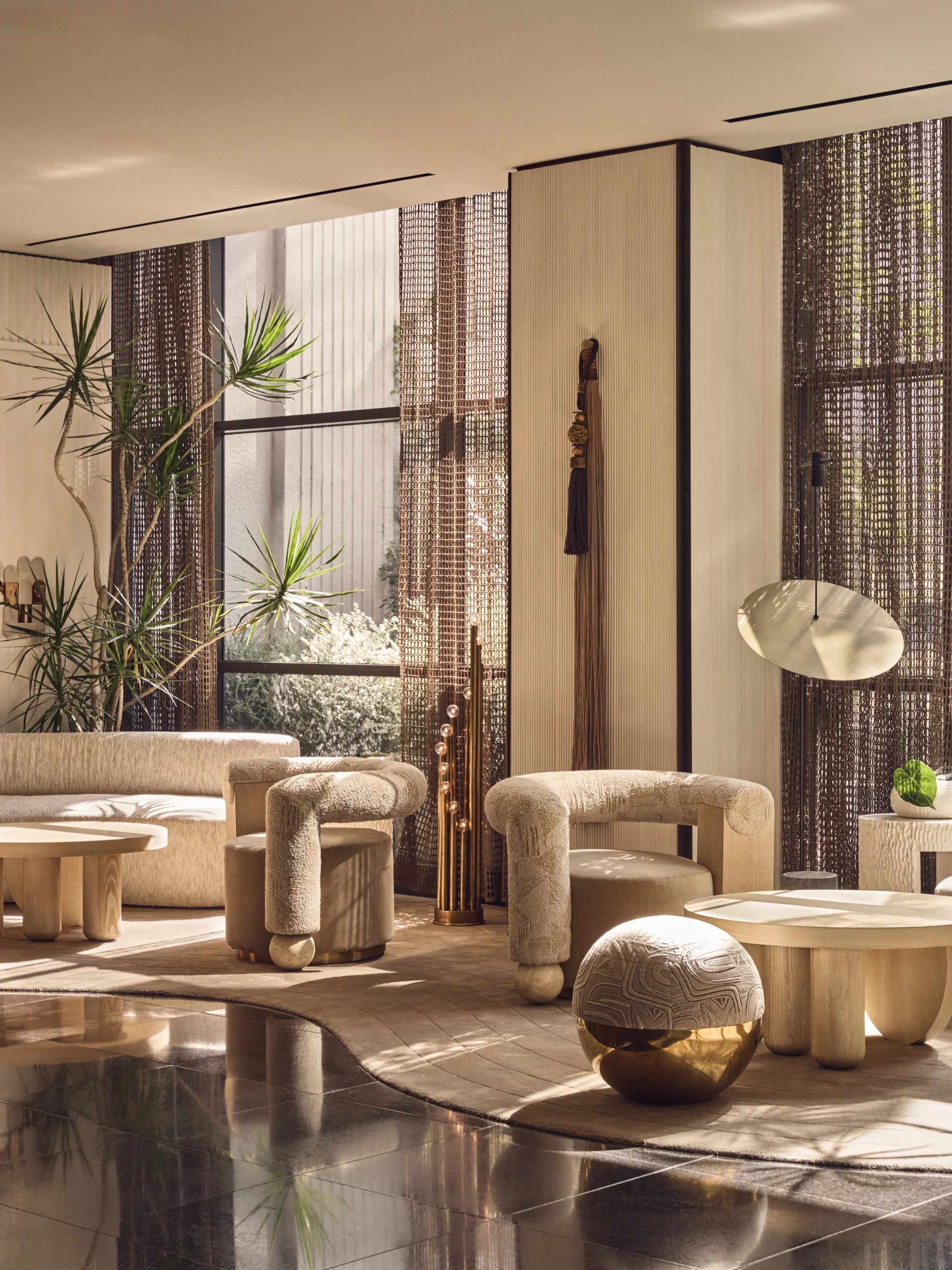 A modern, stylish interior living space with beige upholstered furniture, round ottomans, wooden tables, a potted plant, and large windows with patterned curtains allowing natural sunlight.