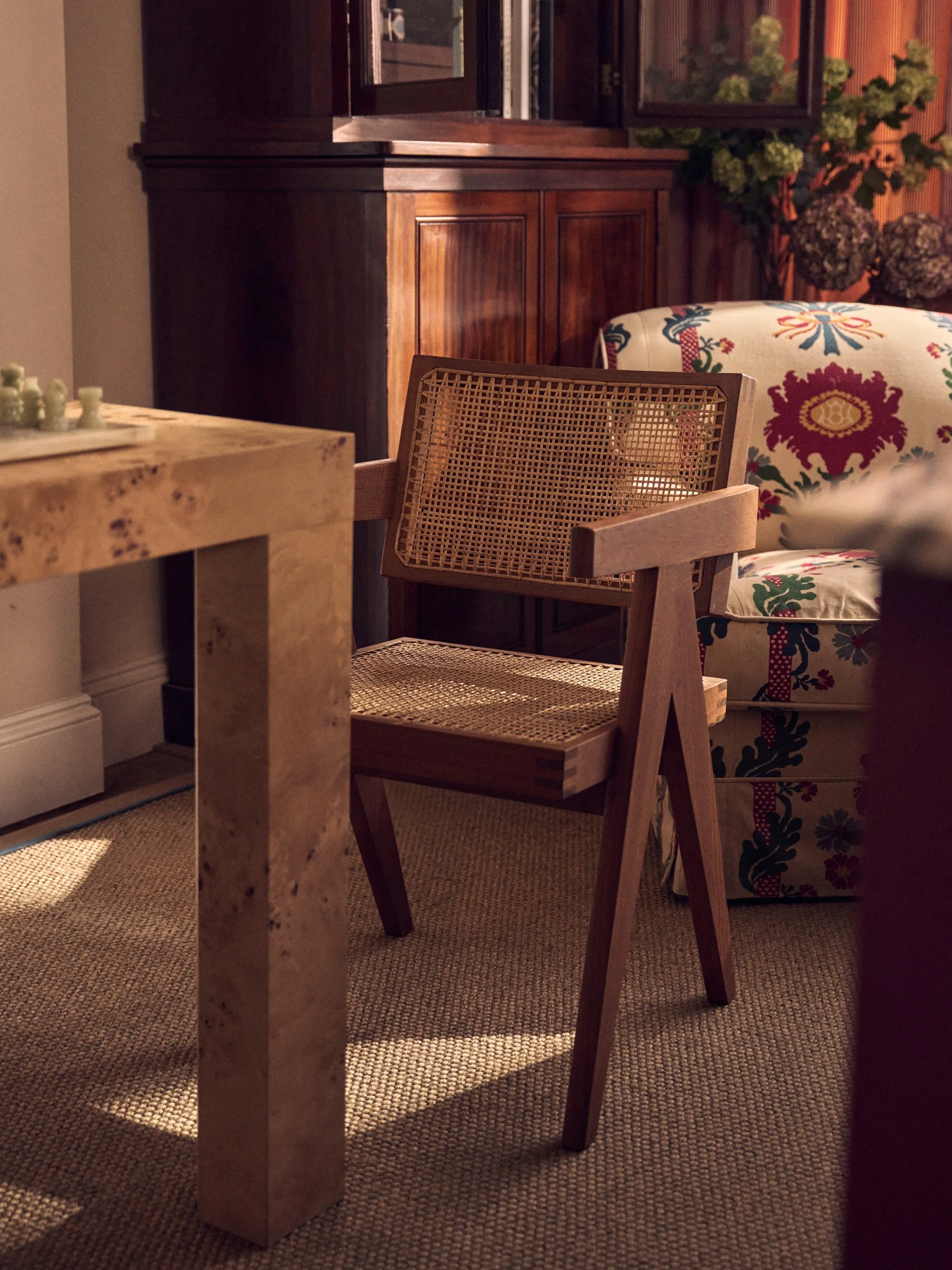 A wooden dining chair with cane seat and backrest in front of a patterned upholstered chair, a wooded china cabinet, and a textured beige carpet.