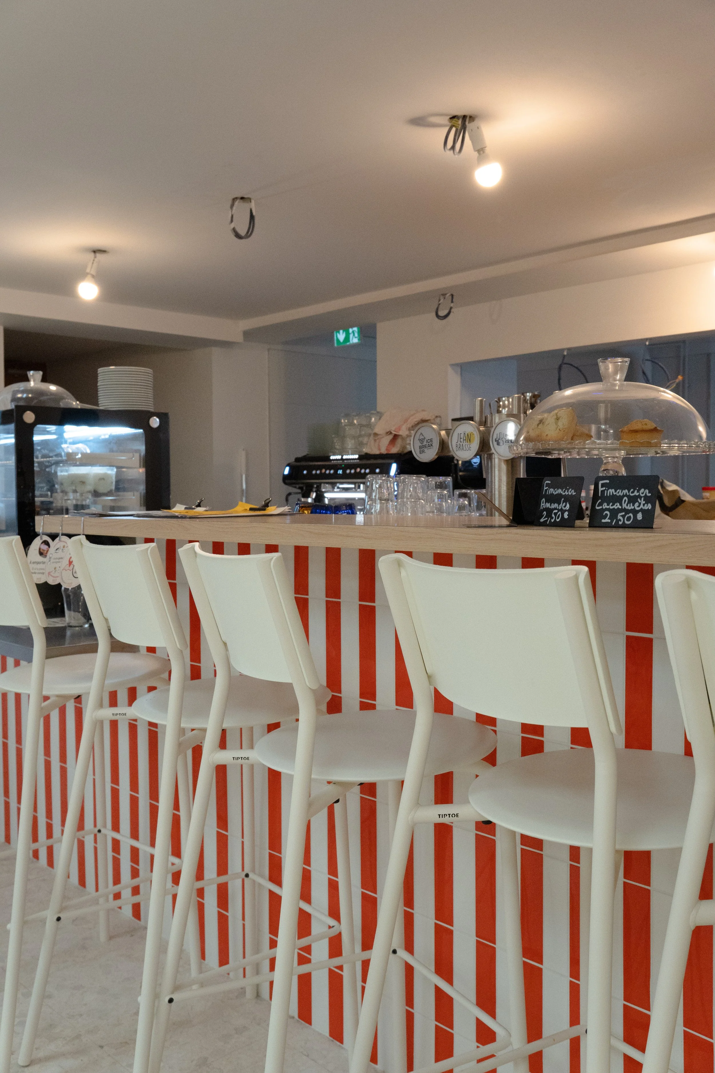 Intérieur d'un café avec un comptoir en bois, plusieurs chaises, décor en bois sur les murs, étagères avec bouteilles et verres, menu affiché, et cookies sur le comptoir.