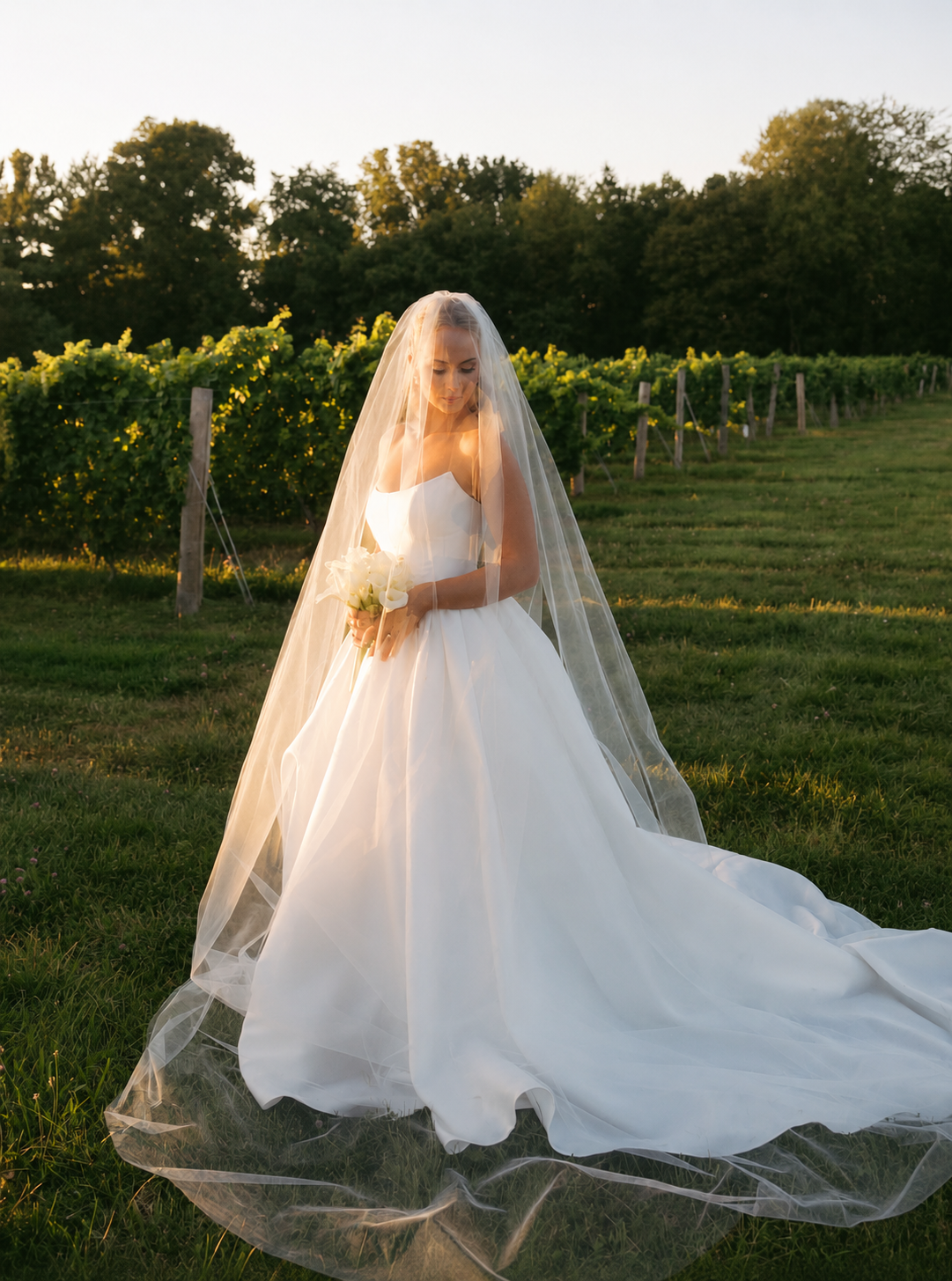 A bride in a white wedding gown and veil holding a bouquet in a field at sunset.