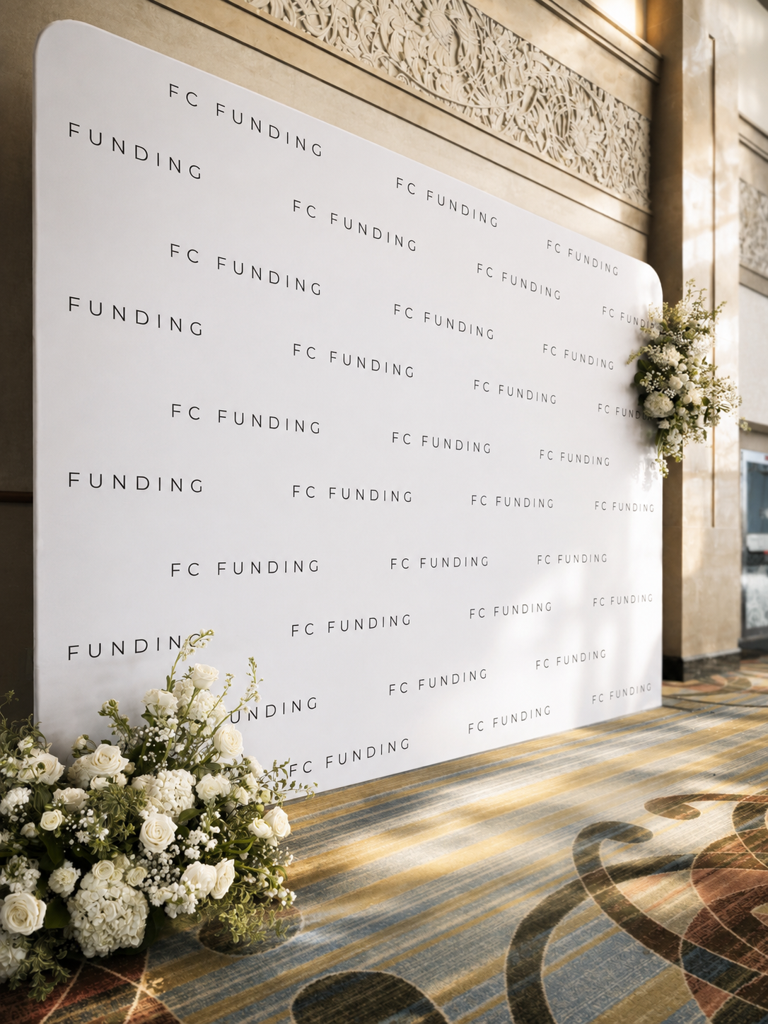 A white backdrop with black text repeating the words "FC FUNDING" and "FUNDING" in a diagonal pattern, decorated with white floral arrangements on the top right and bottom left corners, set against a patterned carpeted floor and ornate interior wall.