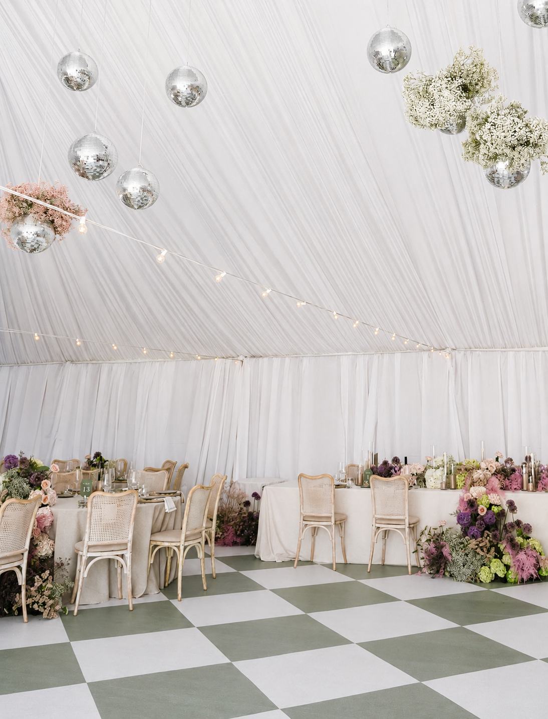 Indoor event space with white draped ceiling, disco balls, string lights, decorated tables with chairs, and floral arrangements on and around the tables.