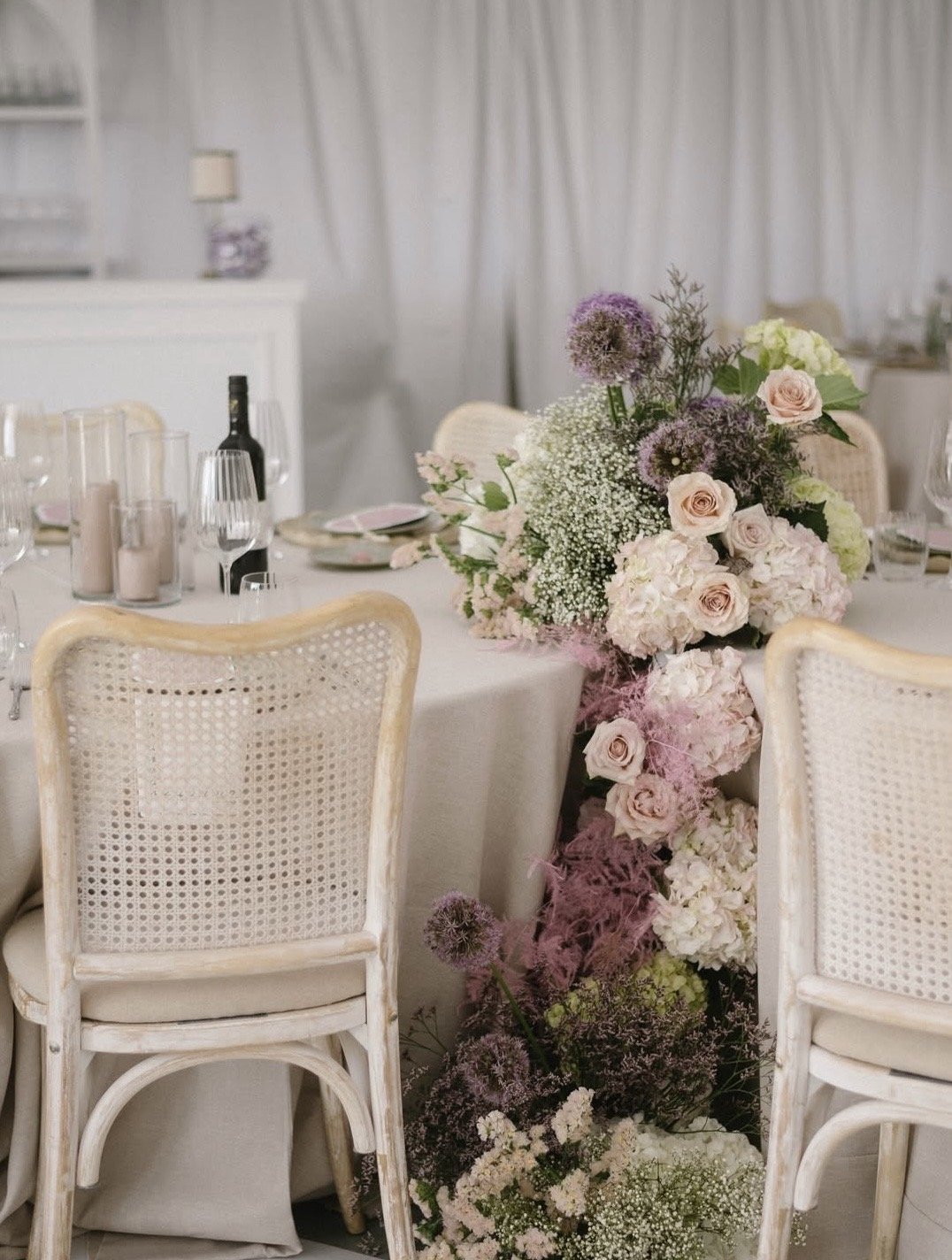 Elegant dining room table with a large pastel flower arrangement featuring roses, hydrangeas, and baby's breath, surrounded by white chairs, set for a formal event.