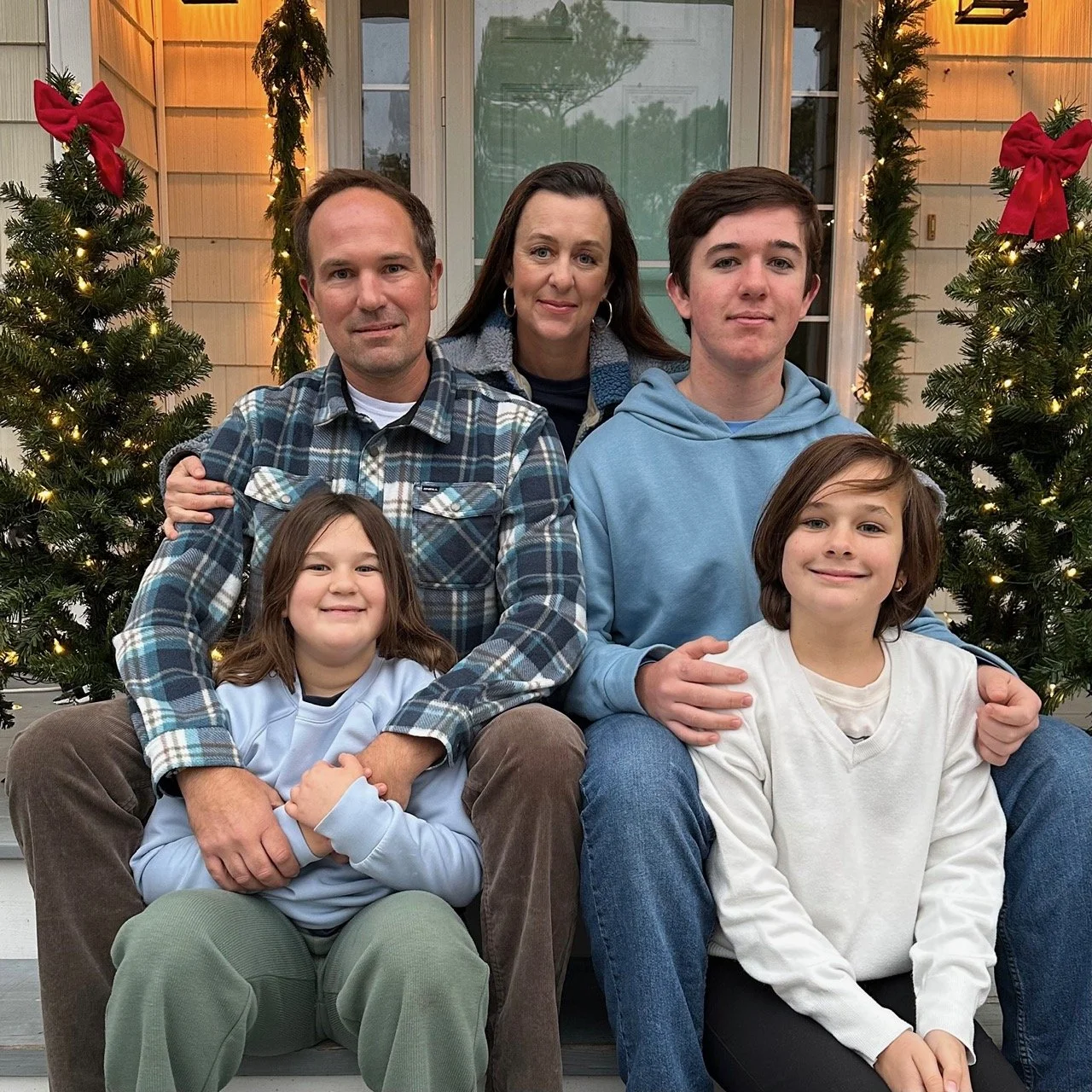 A family of five sitting on a porch decorated with Christmas trees and greenery with red bows, during the holiday season. This picture is of the family who owns Trusty Sail and Paddle from year 2025.