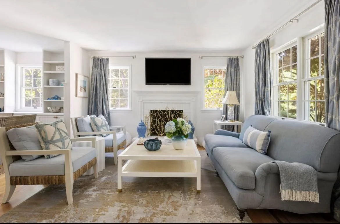 Bright living room with white walls, large windows, gray sofa, and matching armchairs with light-colored cushions, coffee table with decorative vases and flowers, fireplace, TV mounted above, and curtains.