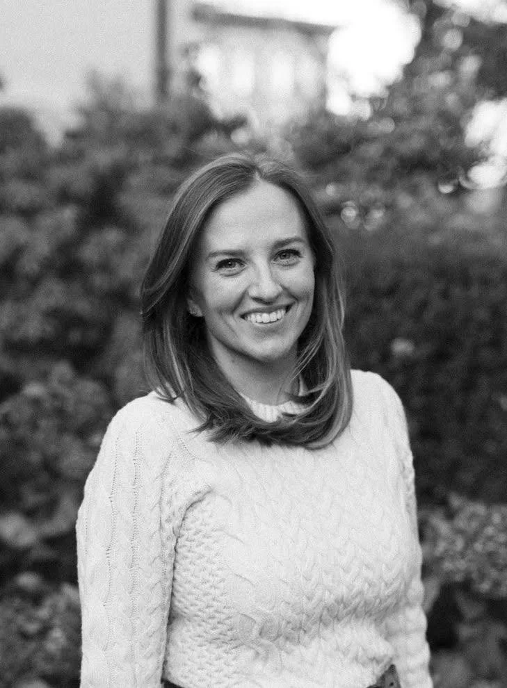 A smiling woman with shoulder-length hair wearing a knitted sweater outdoors with blurred trees in the background.