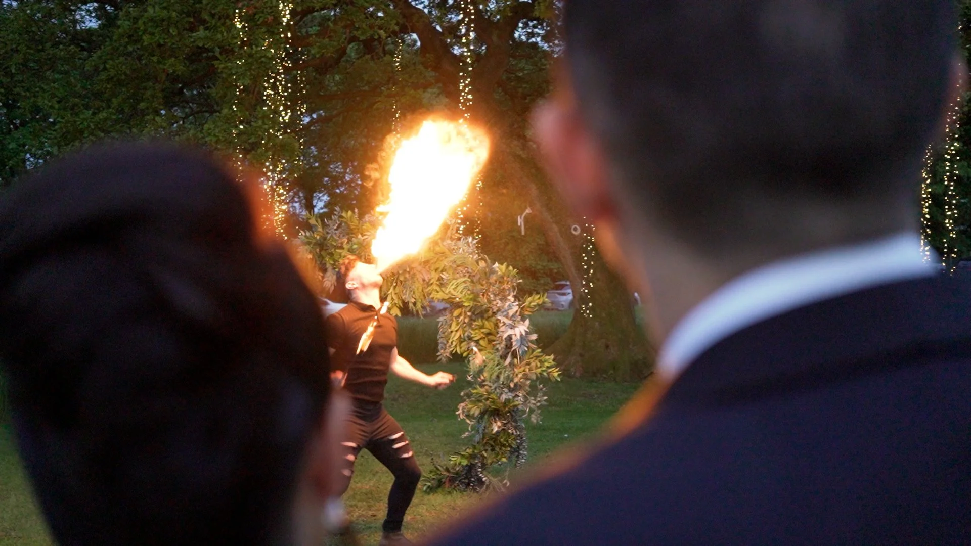 A fire breather performs at dusk in the gardens at The Tipis at Riley Green, filmed over the shoulders of the bride and groom with fairy lights glowing in the trees behind