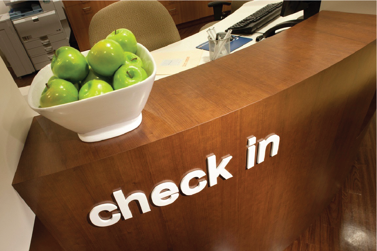 Reception desk with a "check in" sign and a bowl of green apples, office setting.