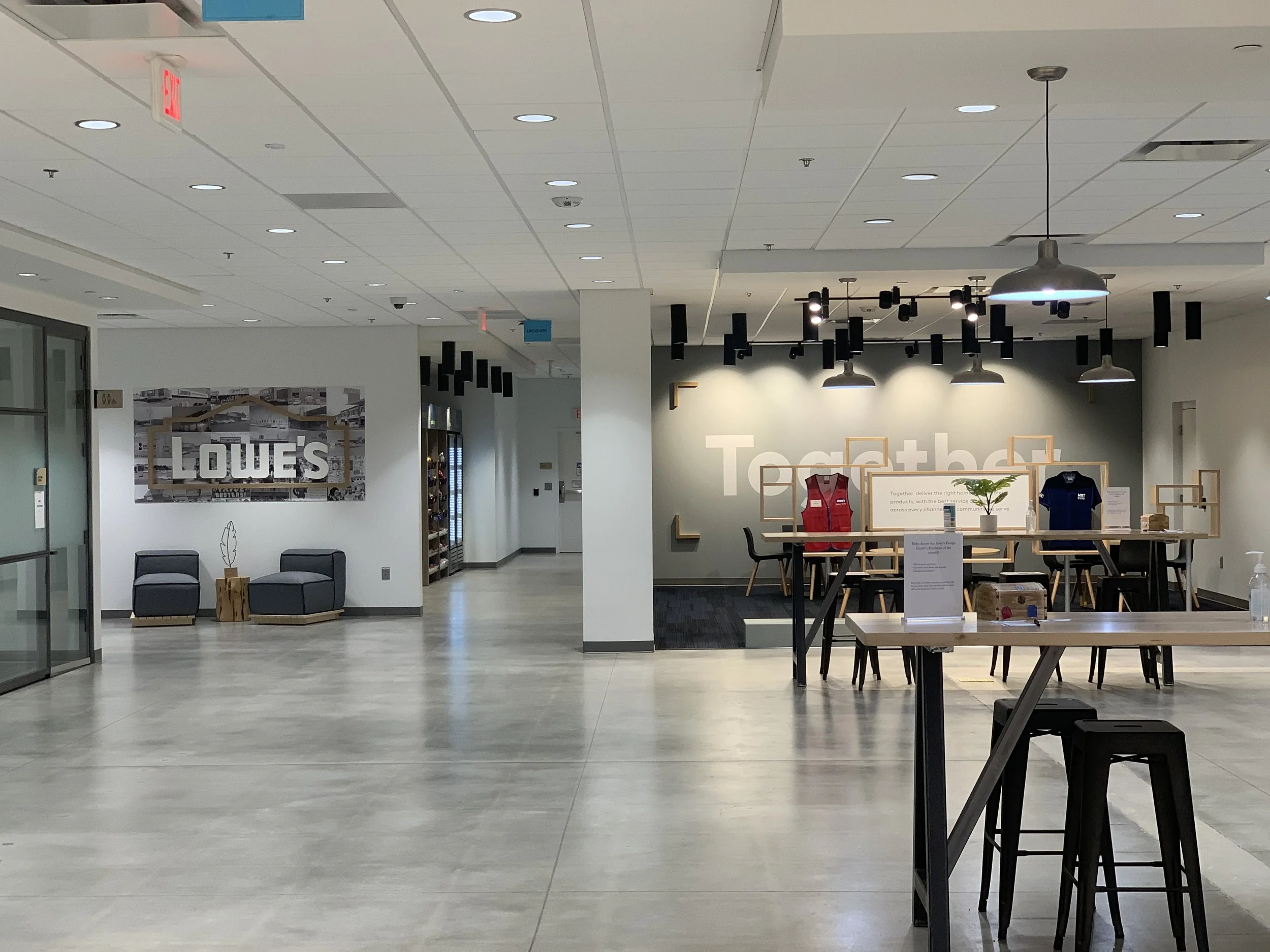Open office space with a modern design, featuring tables, chairs, and apparel on display. A wall displays a large sign that reads 'LOWE'S.' Overhead lighting fixtures hang from the ceiling, and a small lounge area is visible to the left.