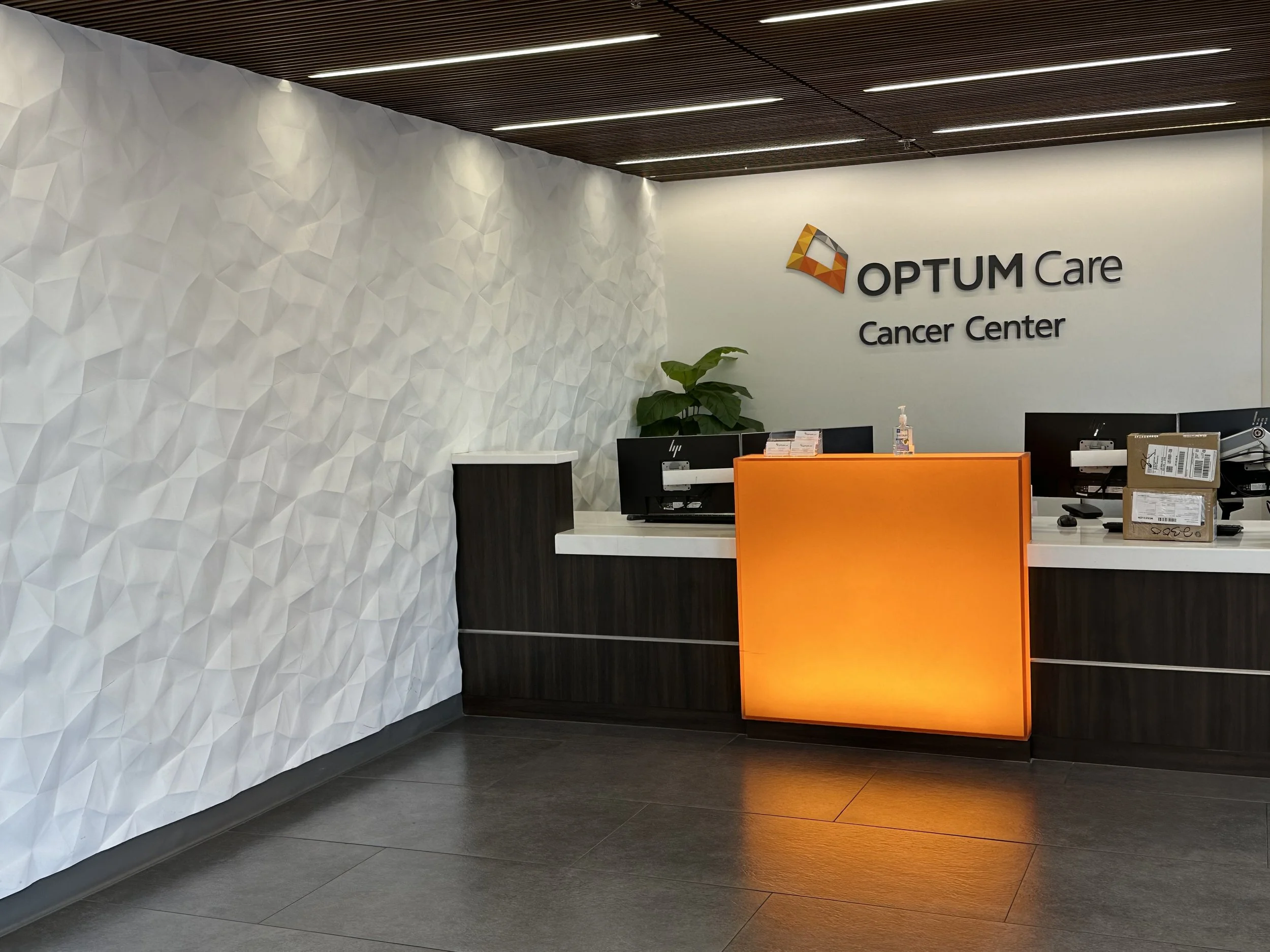 Optum Care Cancer Center reception desk with textured wall, orange-lit counter, and computer stations.