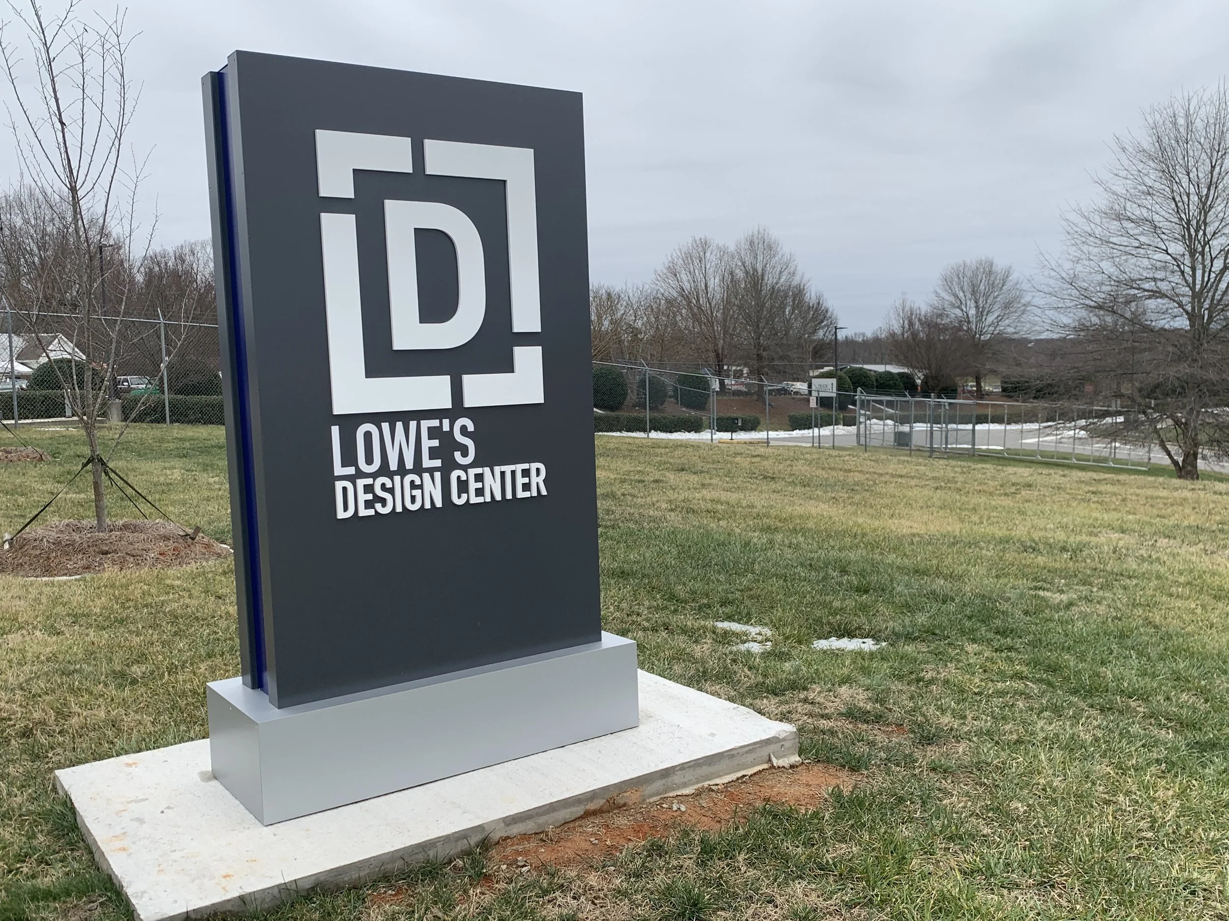 Outdoor sign for Lowe's Design Center on a grassy lawn with trees and overcast sky in the background.