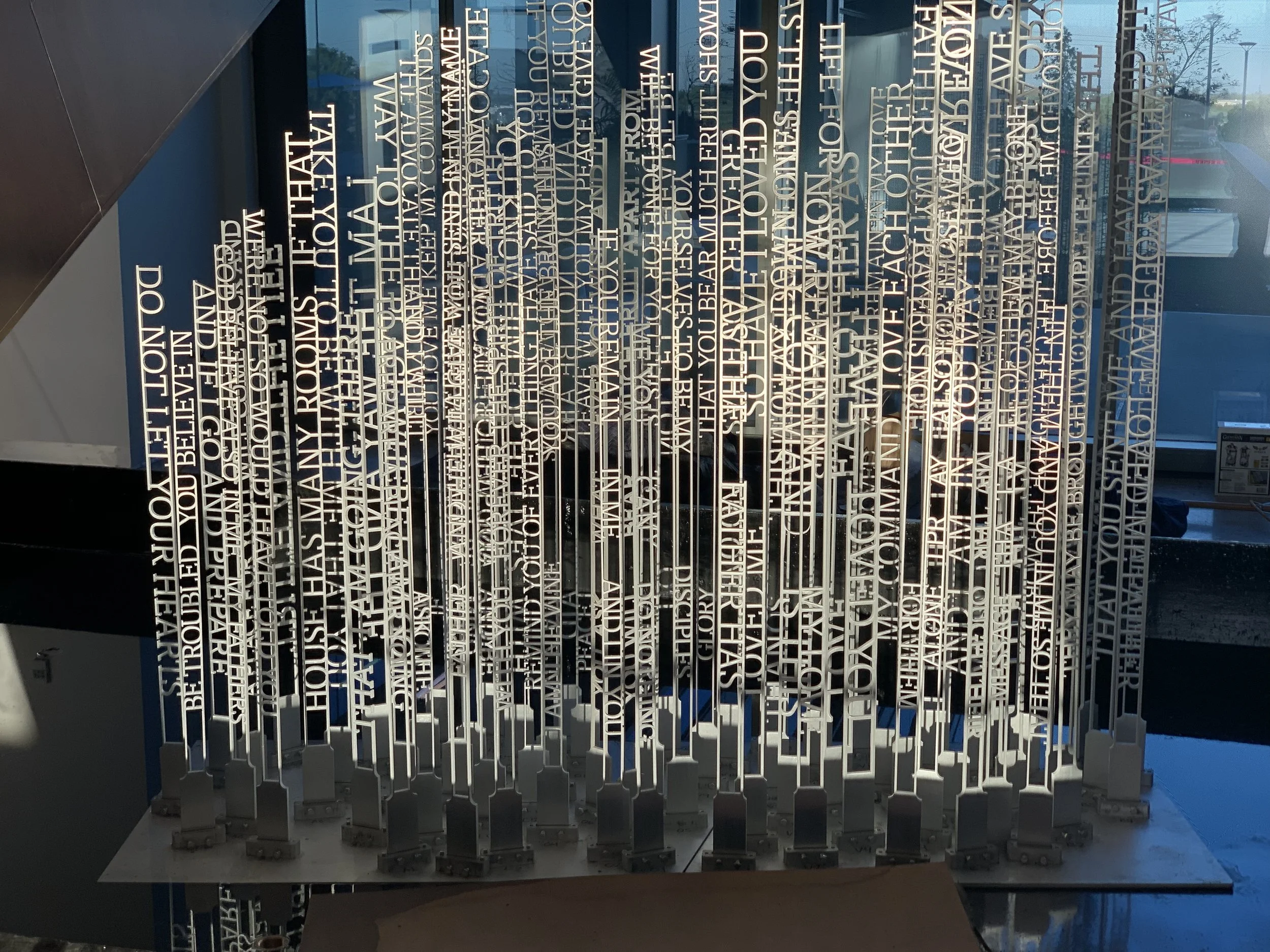 Sculpture of vertical metal text strips on stands, displaying various words and phrases in a modern design. Background window showing outdoor view.