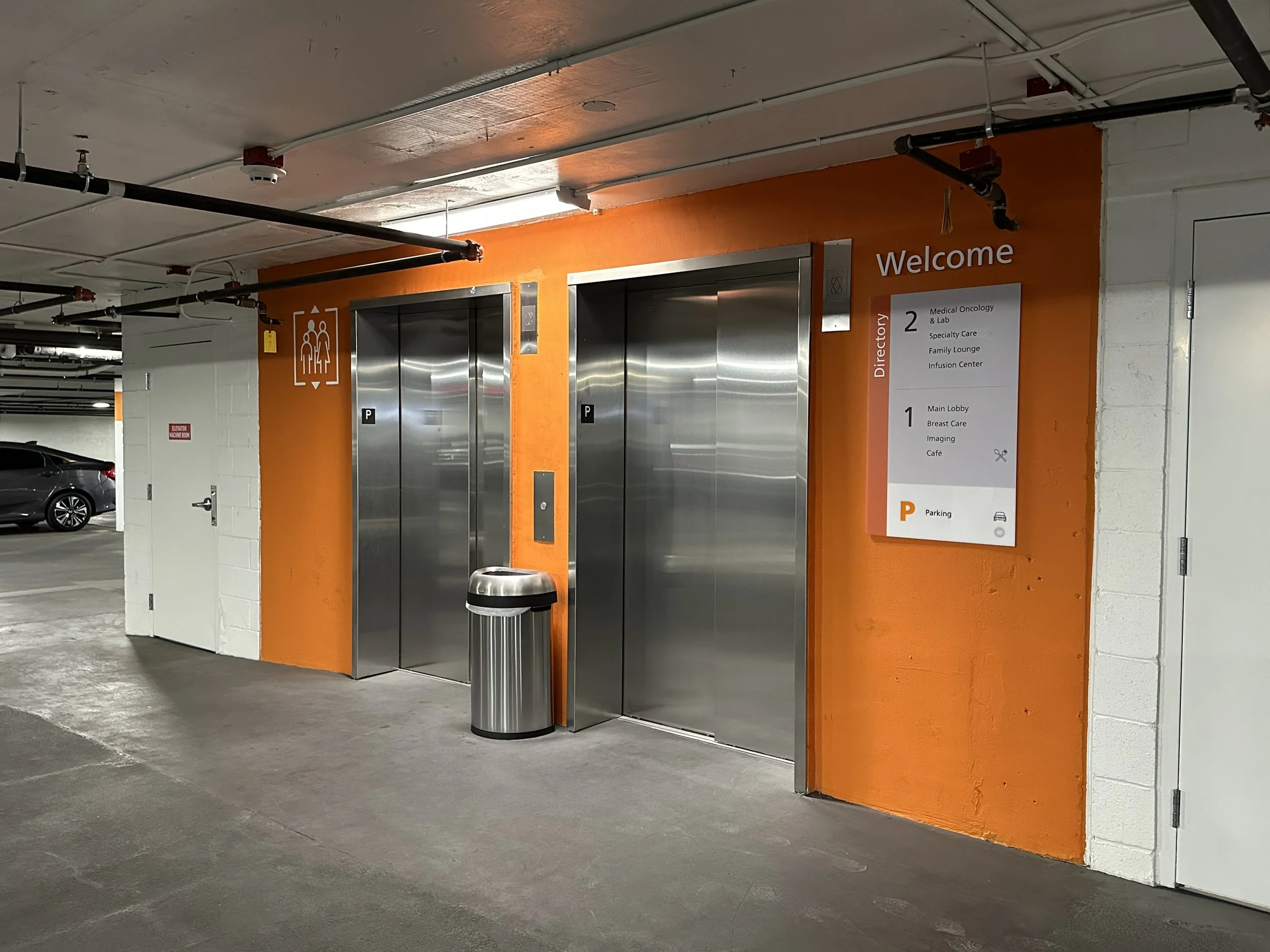 Parking garage with two elevators, an orange wall, a sign with directory information, and a trash bin.