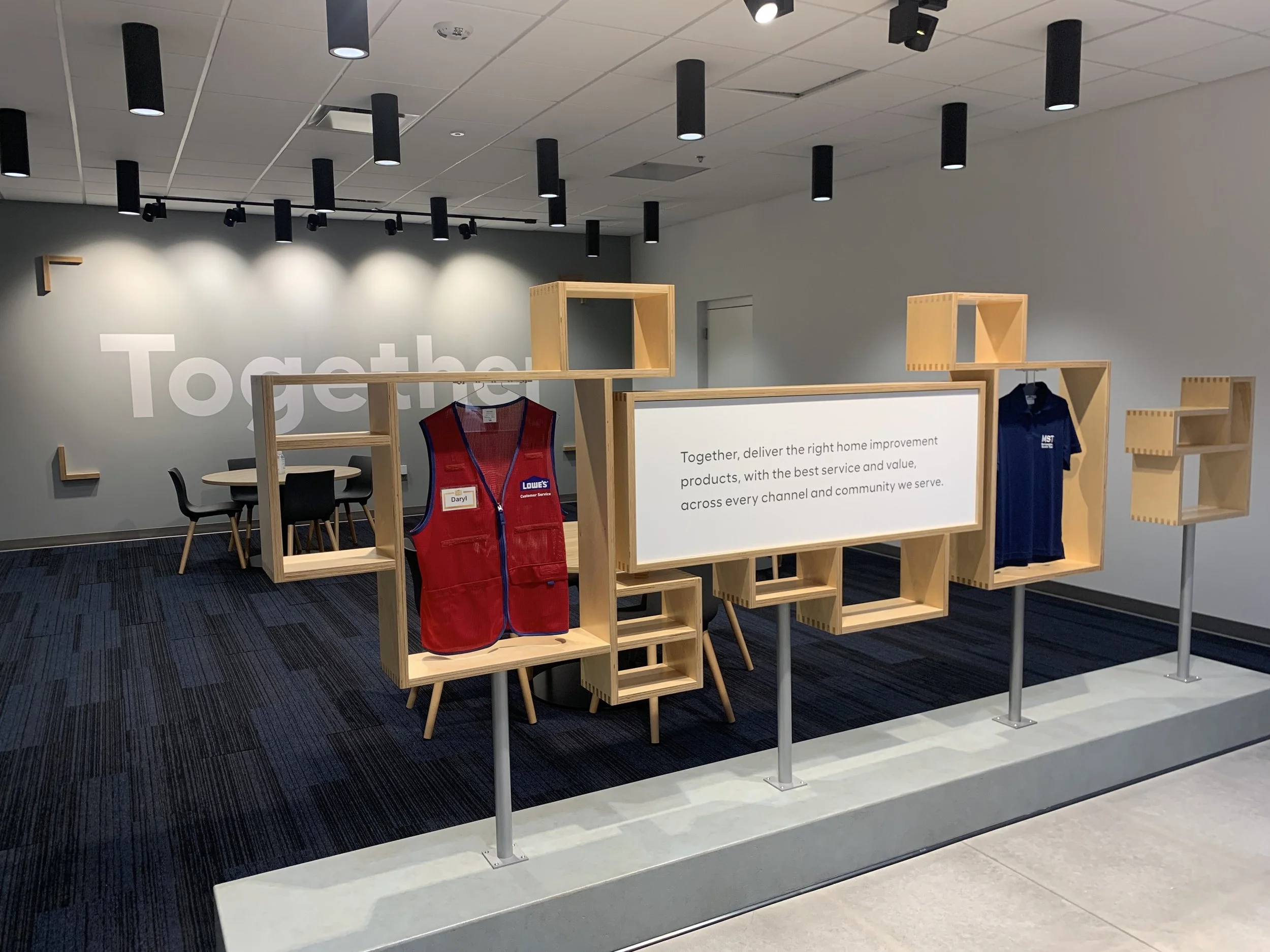 Retail store showroom with modern shelving, a red vest, and a blue shirt displayed. Wall includes the word "Together" and a mission statement about delivering home improvement products. Meeting area in the background with a round table and chairs.