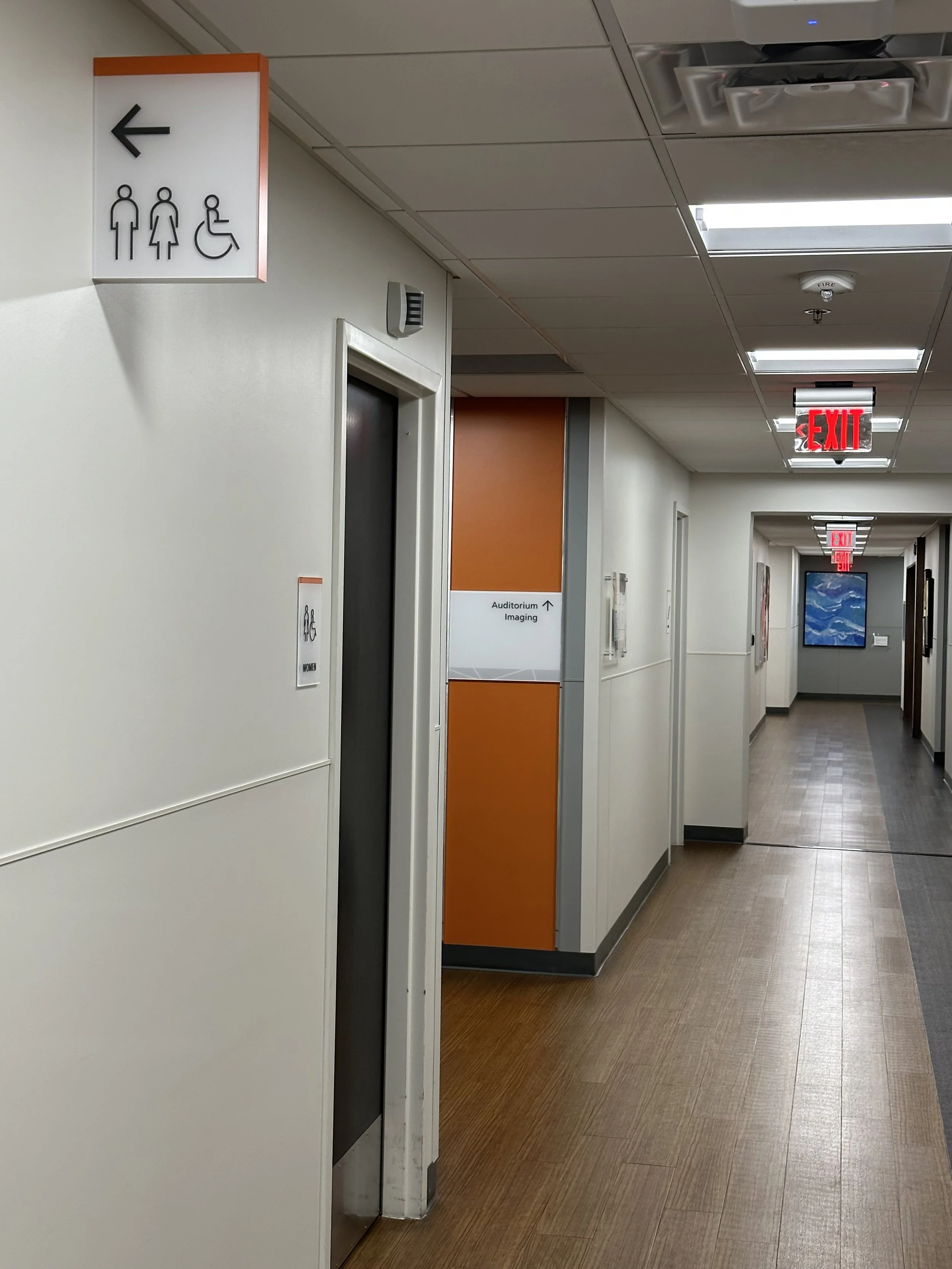 A hallway in a building with wood-patterned flooring and white walls. On the left, there is a restroom sign indicating male, female, and accessible facilities. The hallway continues with "Auditorium" and "Imaging" directions shown, and an exit sign o