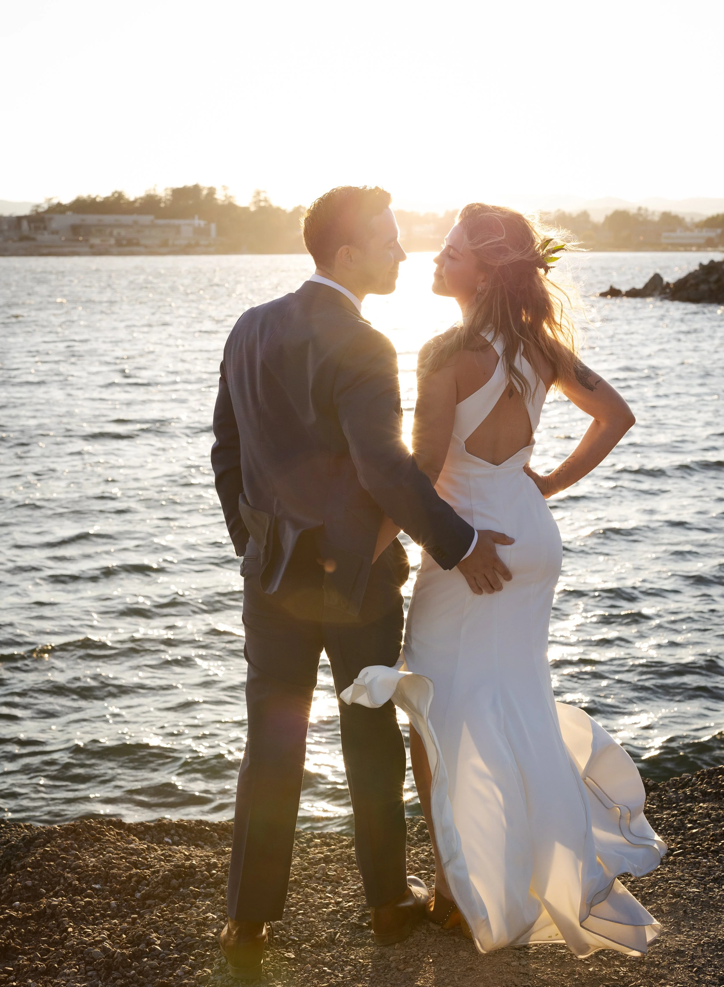 A couple in wedding attire standing by the water at sunset, facing each other and holding hands.