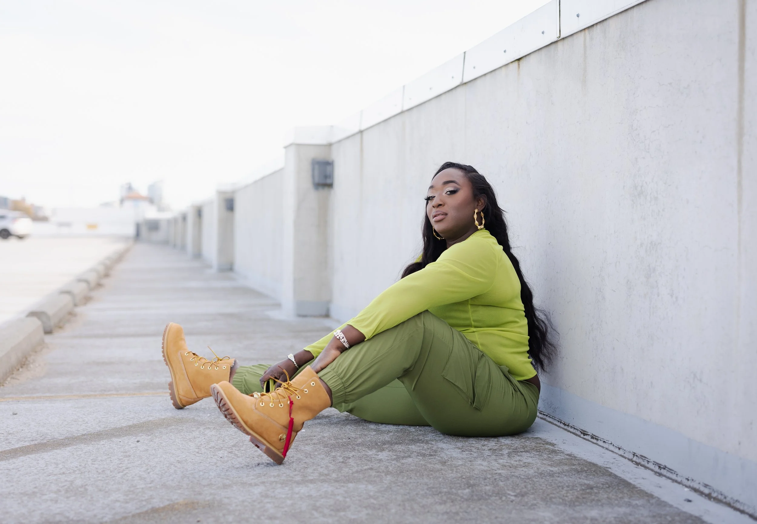 A woman sitting on the ground against a concrete wall, wearing a lime green top, cargo pants, and yellow boots, with long dark hair and gold jewelry.