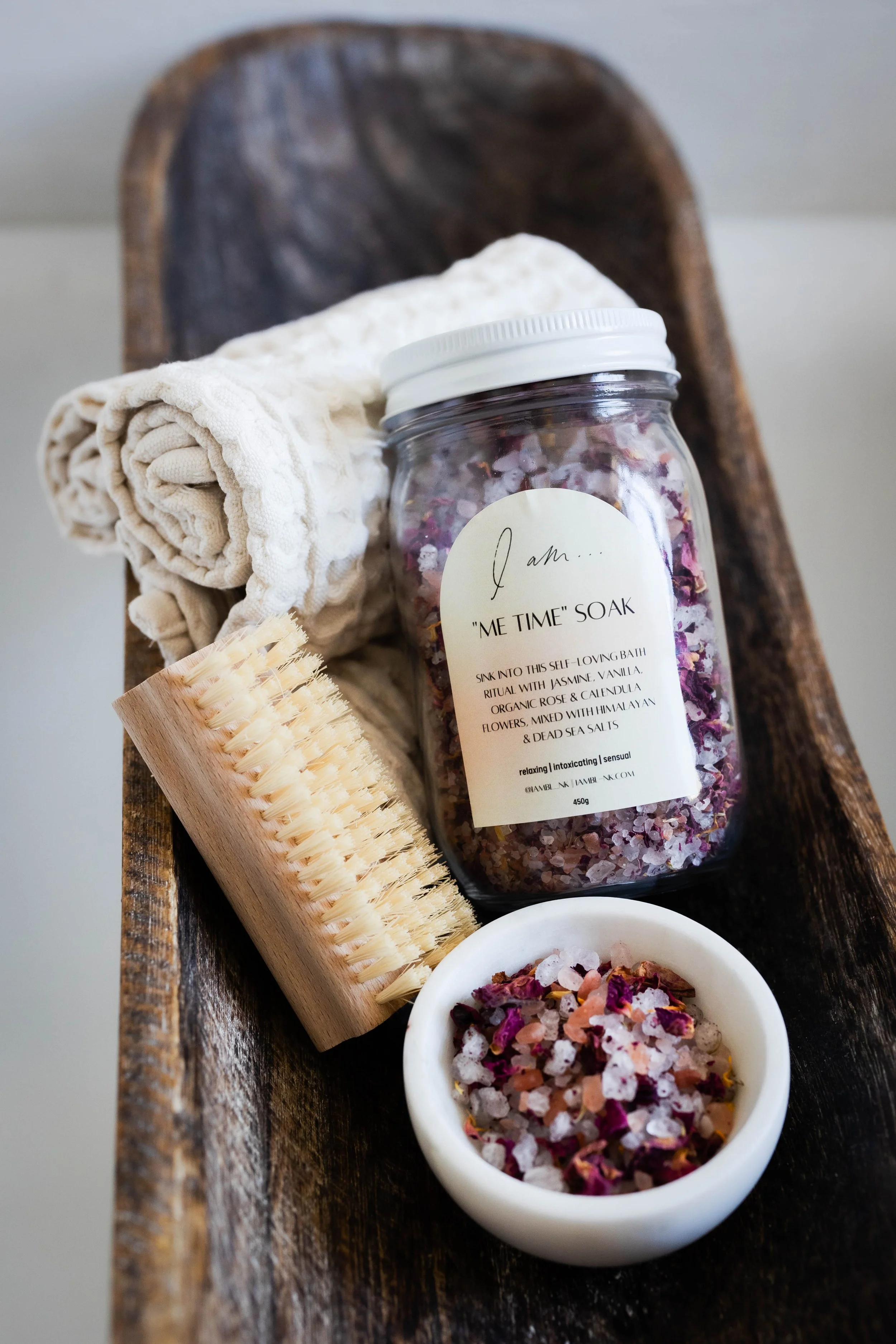 A jar of bath soak labeled 'Me Time Soak' with dried flowers, a wooden bath brush, a rolled towel, and a small bowl of bath salts on a wooden tray.