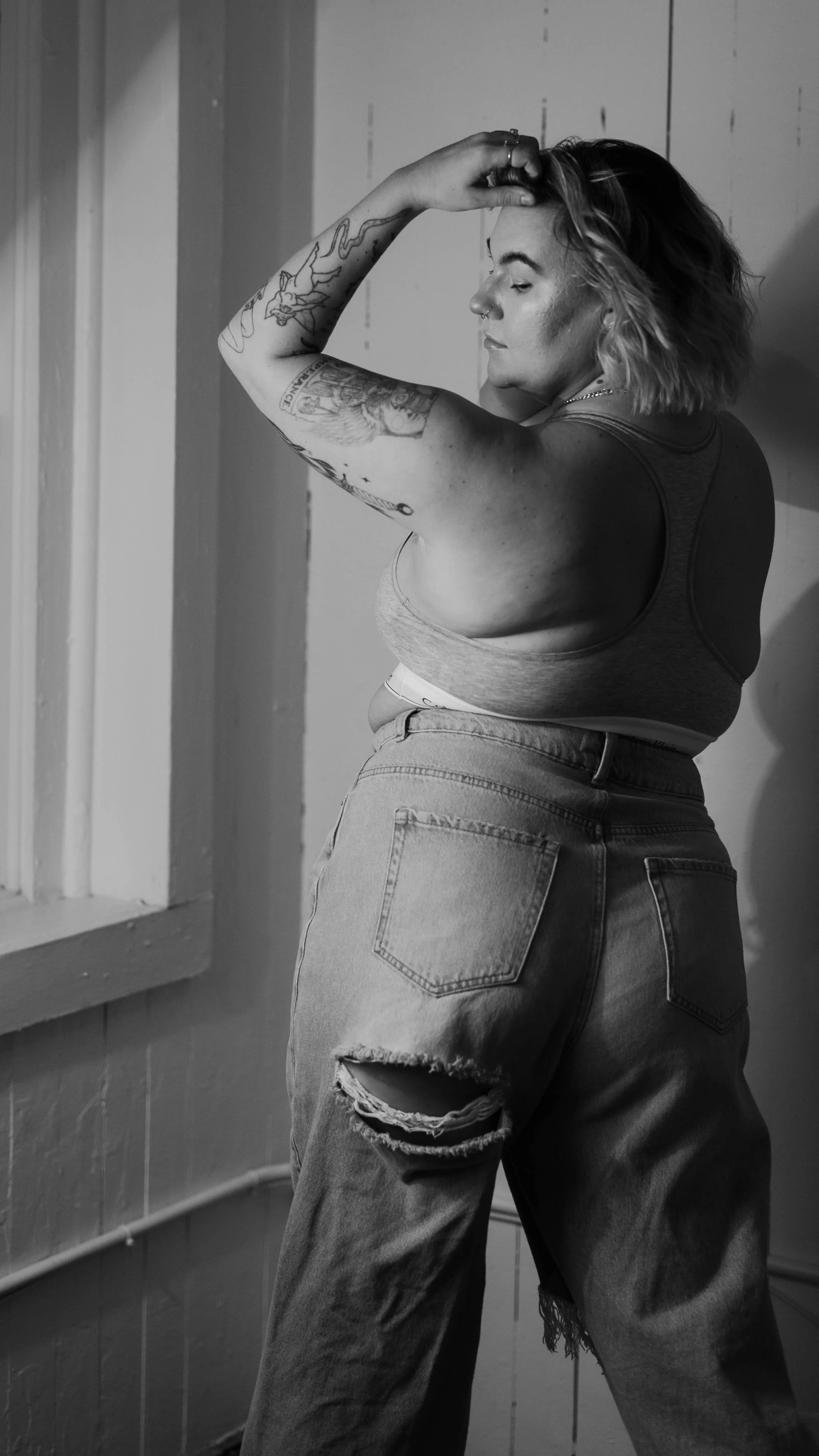 A woman with tattoos on her left arm, styled in a tank top and ripped jeans, poses with her hand on her head near a window in a black and white photo.