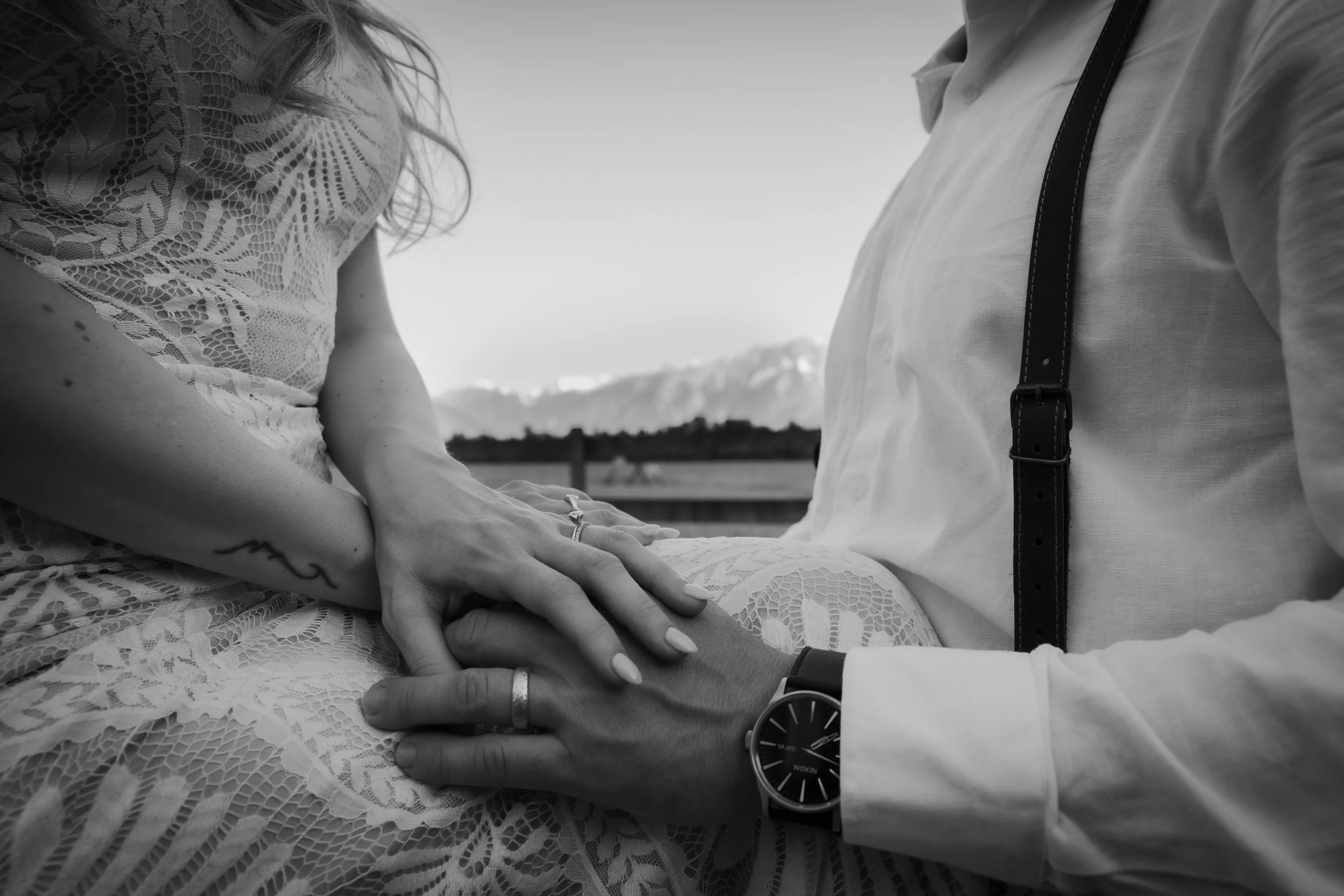 A black and white close-up photo of a couple, with the woman sitting on the man's lap, holding hands and showing wedding rings. The woman is wearing a lace dress and has a tattoo on her arm. The man wears a watch, a white shirt, and suspenders. In the background, there are mountains and a fence, suggesting an outdoor setting.