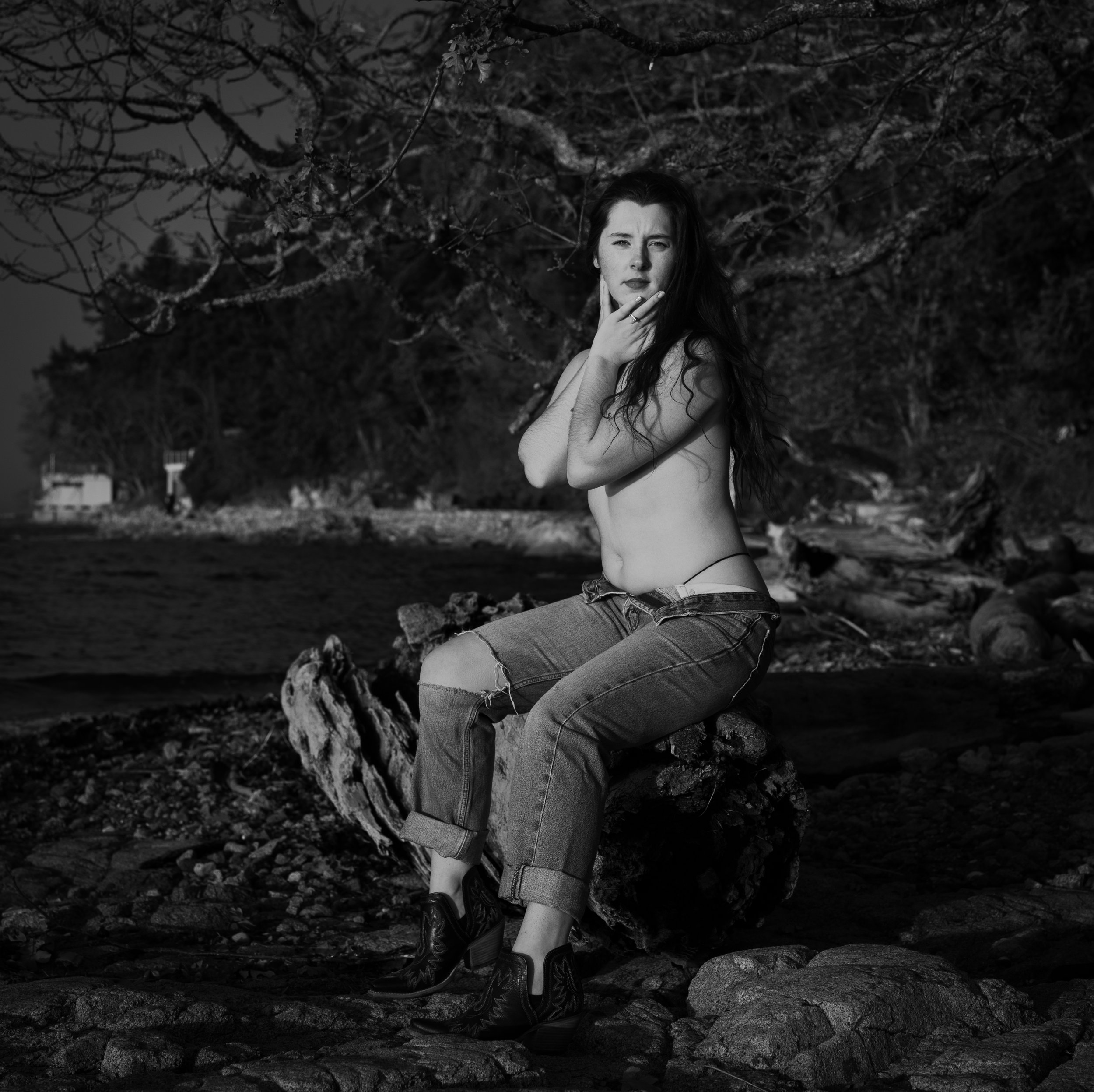 A black and white photo of a woman sitting on a tree stump outdoors, with trees and a shoreline in the background, topless and wearing ripped jeans and high-heeled shoes.