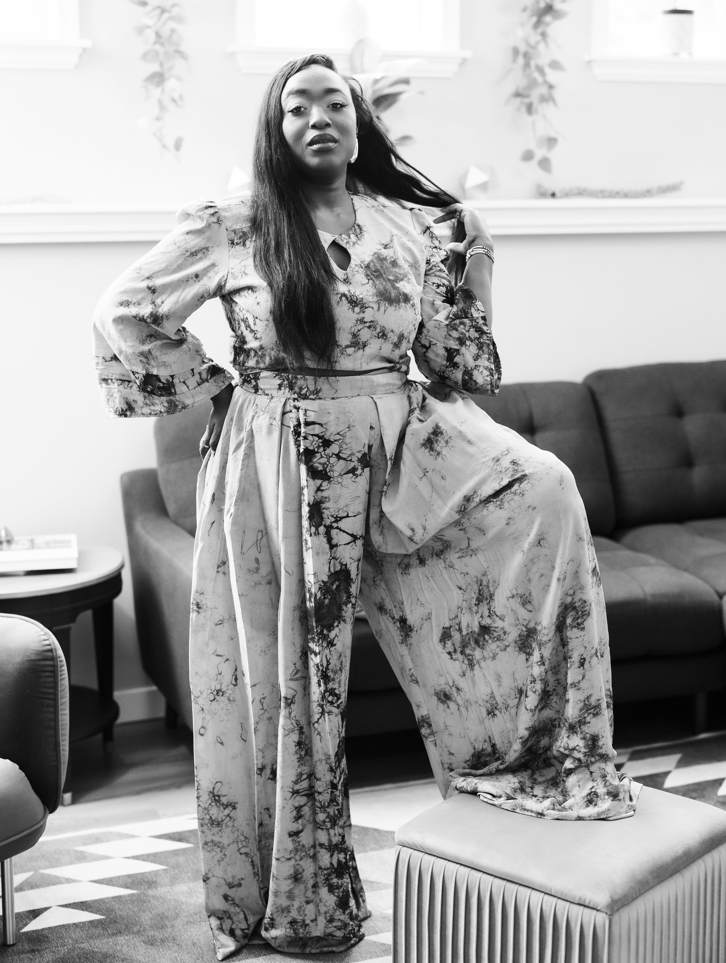 A woman with long hair wearing a tie-dye patterned outfit, standing in a living room with one foot elevated on a stool, and posing confidently.