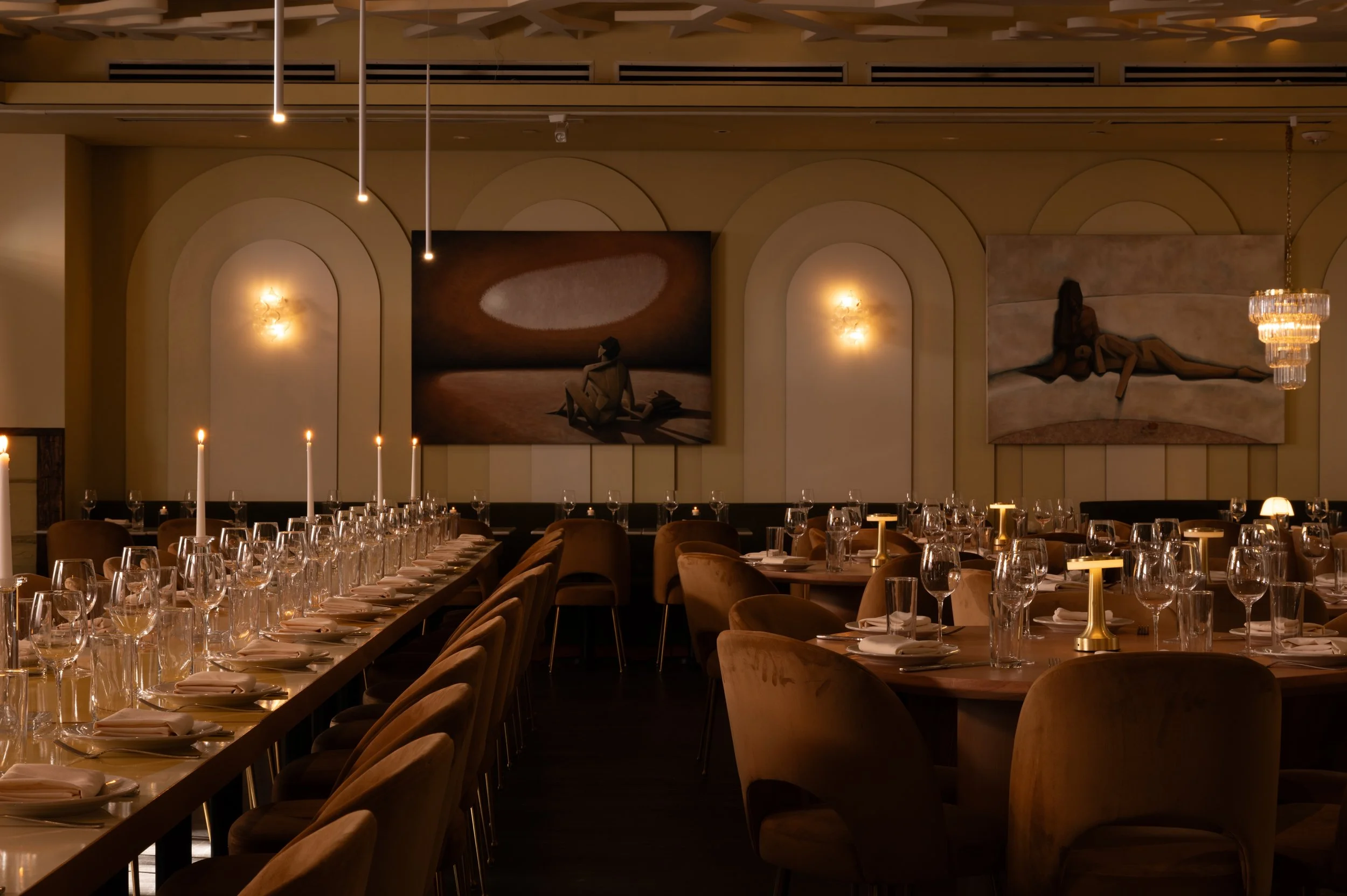 A candlelit dining room with long tables, upholstered chairs, arched walls, and artwork