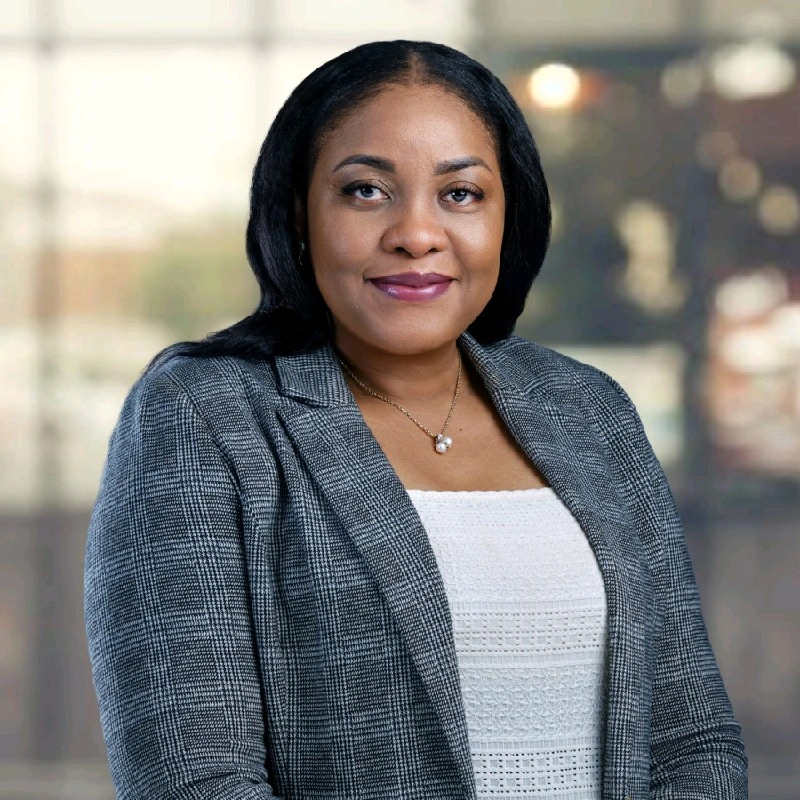 A Black business woman in a navy and white blazer and pearl pendant necklace looks at the camera with a subtle, confident smile