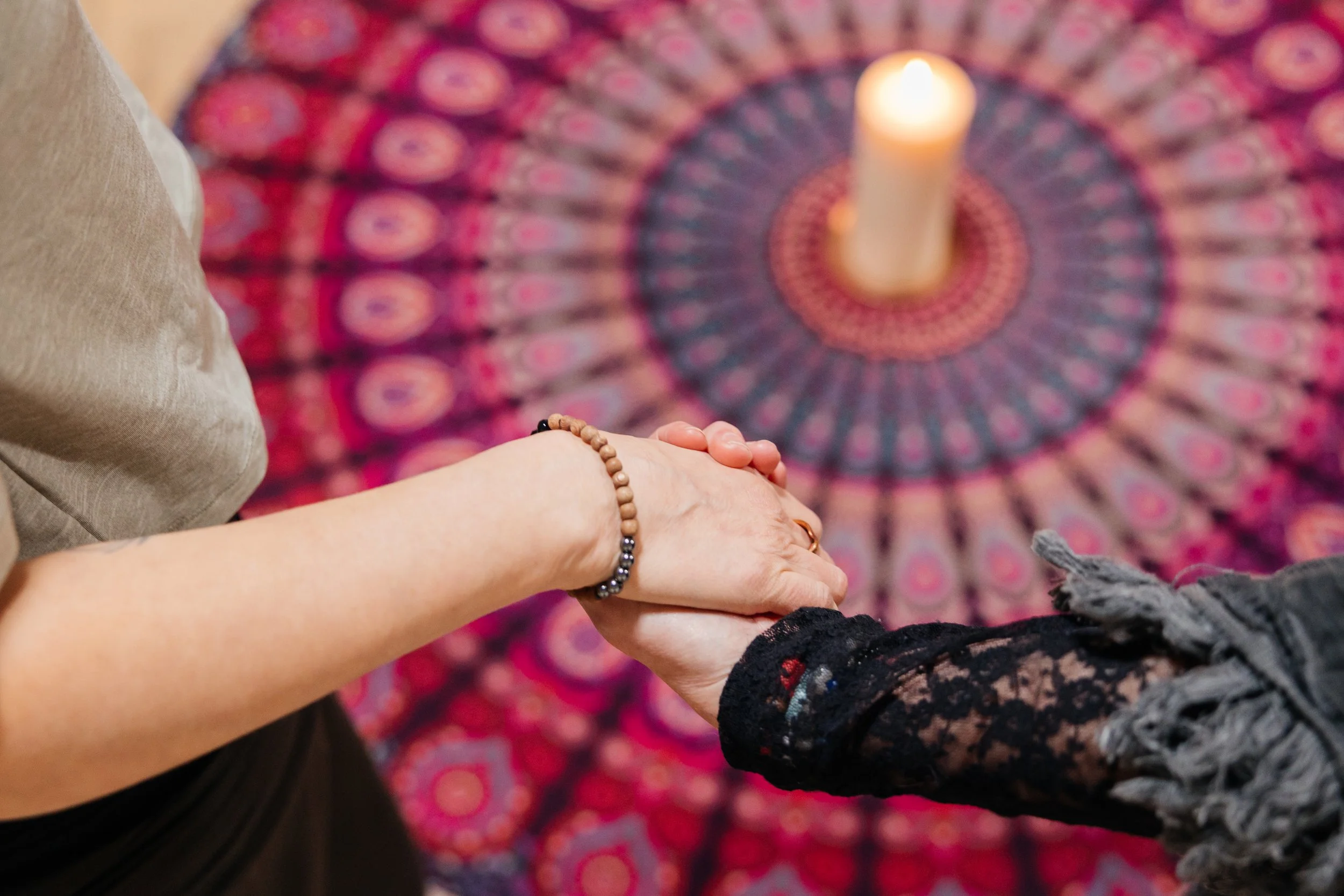 Group of eight women sitting in a circle on mats around a lit candle on a colorful round rug in a room with wooden floor and minimal decor, engaging in a meditation or mindfulness session.