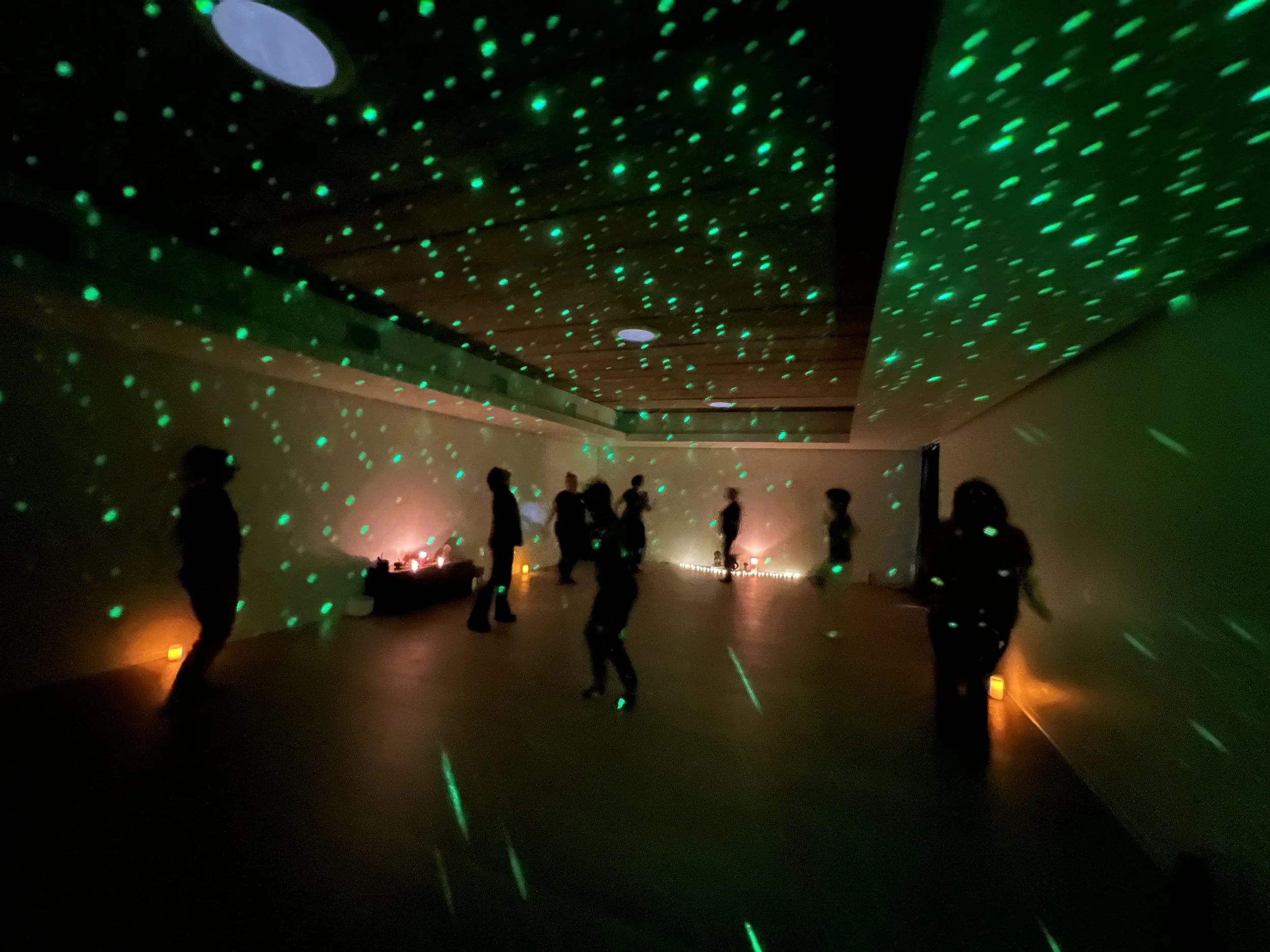 A dark room with green laser light dots projecting onto the ceiling and walls. Several people are standing or walking around, silhouetted against the laser lights.