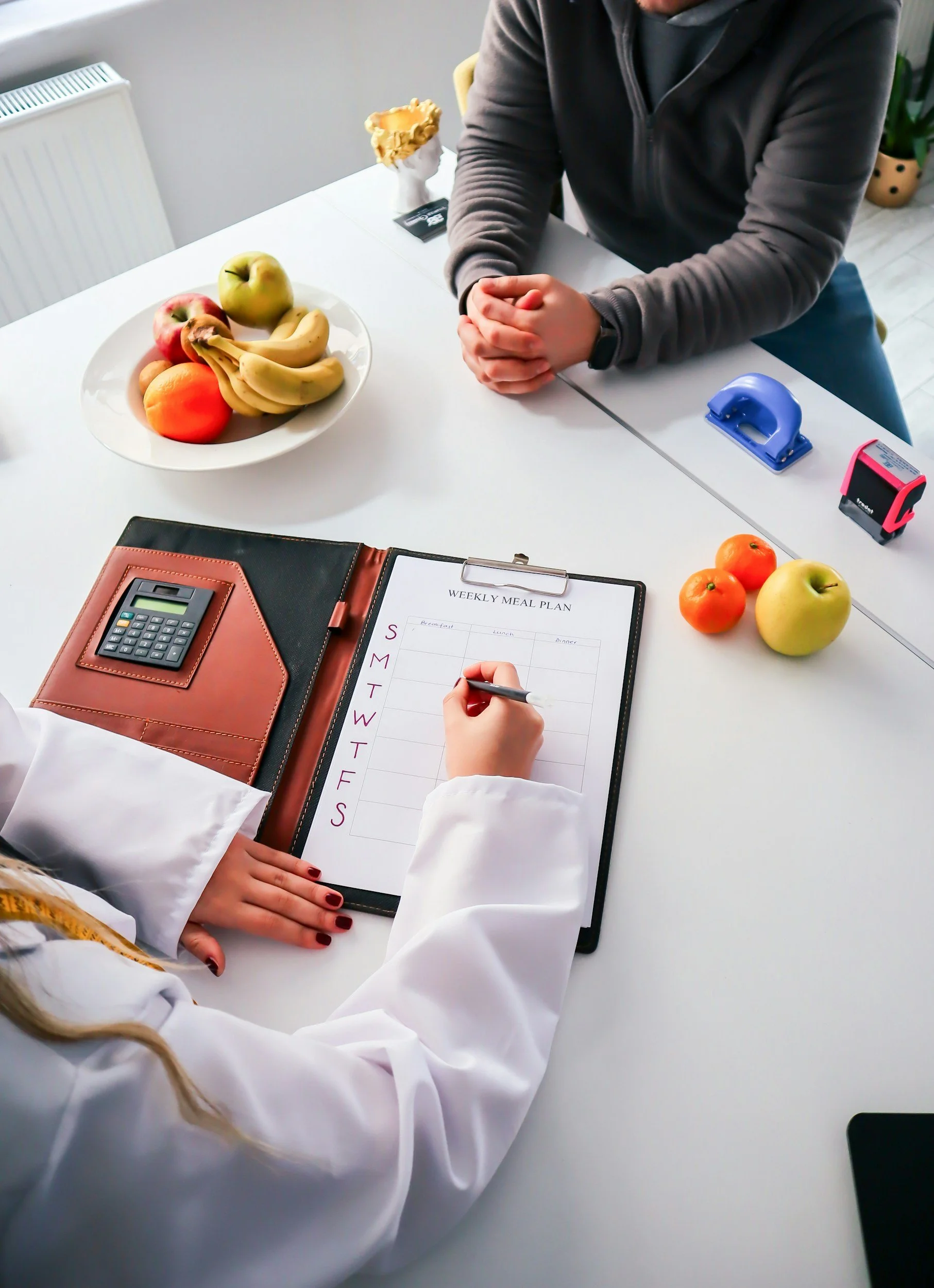 A person with glasses, wearing a gray zip-up jacket, sitting at a white table with a large plate of assorted fruit, including apples, bananas, and a peach. There are also two smaller apples, a peach, and a person writing on a weekly meal plan notepad. Office supplies like a calculator, a brown notebook, and two self-inking stamps are also visible.