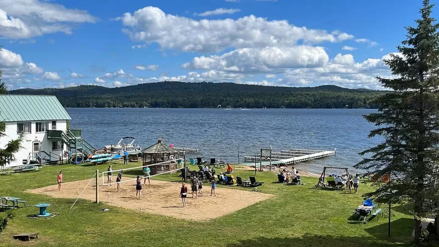 People playing volleyball on a sand court near a lake with dock, boats, and a gazebo, surrounded by green grass, trees, and a building with a green roof, under a blue sky with clouds.