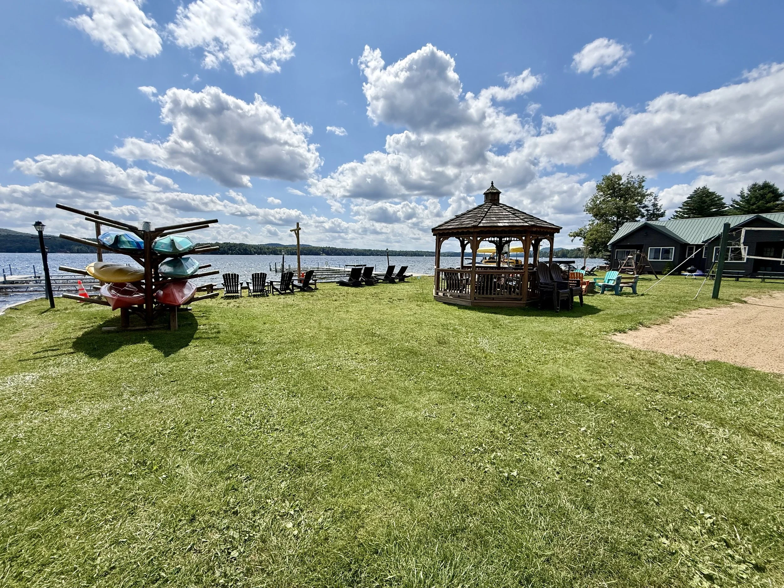 A lakeside scene with a gazebo, Adirondack chairs, and a boat dock, under a partly cloudy sky.
