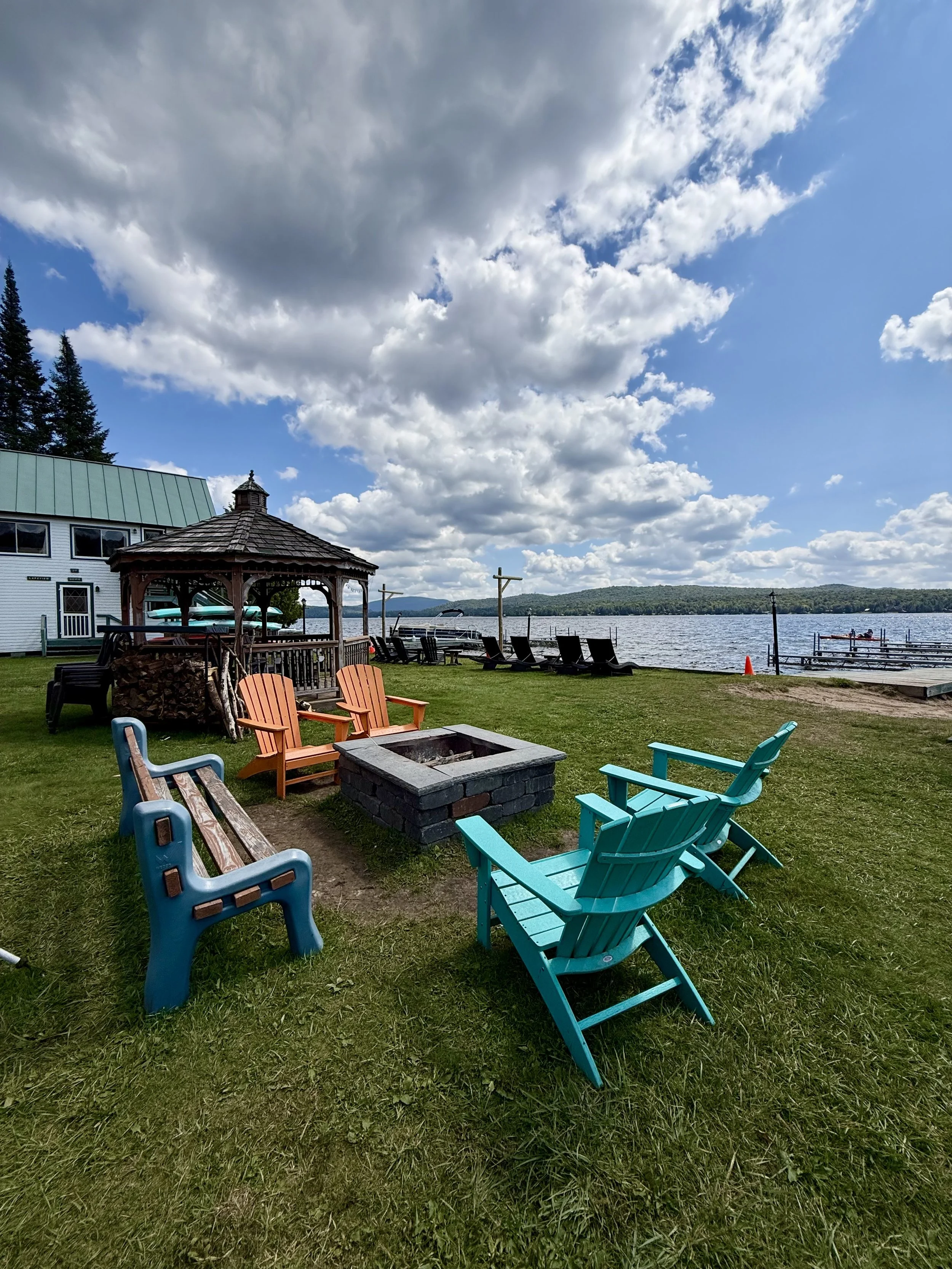 A lakeside outdoor seating area with colorful Adirondack chairs, a fire pit, and a gazebo. In the background, there is a dock with boats and a large body of water under a partly cloudy sky.